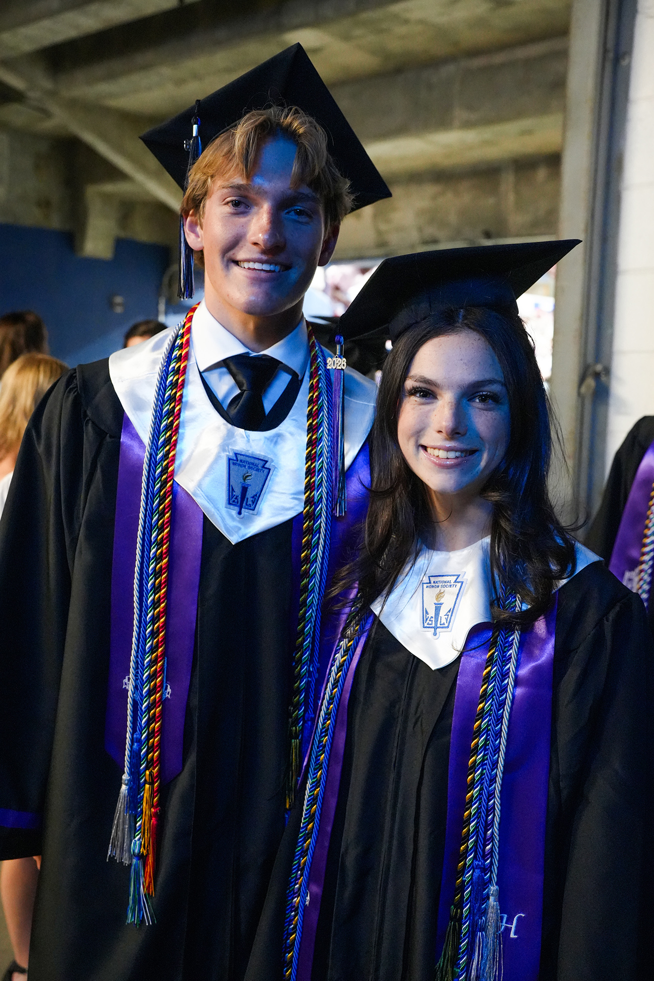 Two individuals in graduation gowns and caps stand close together, smiling. The male wears a tie and a sash with colorful cords. The female wears a sash.