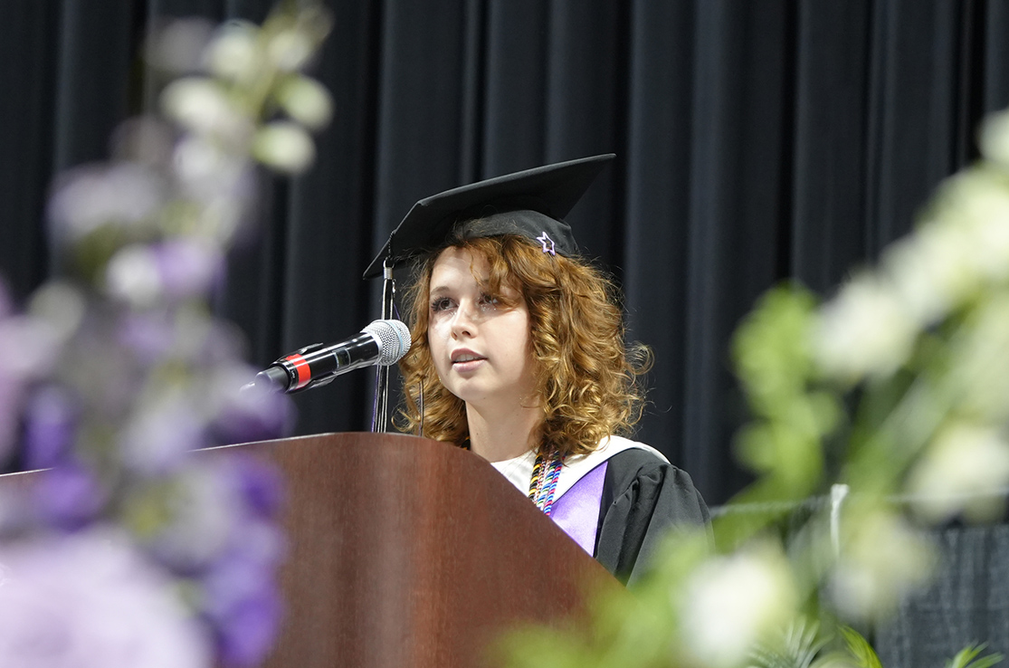 A young person with red hair wears a graduation gown and cap. They speak into a microphone at a podium.