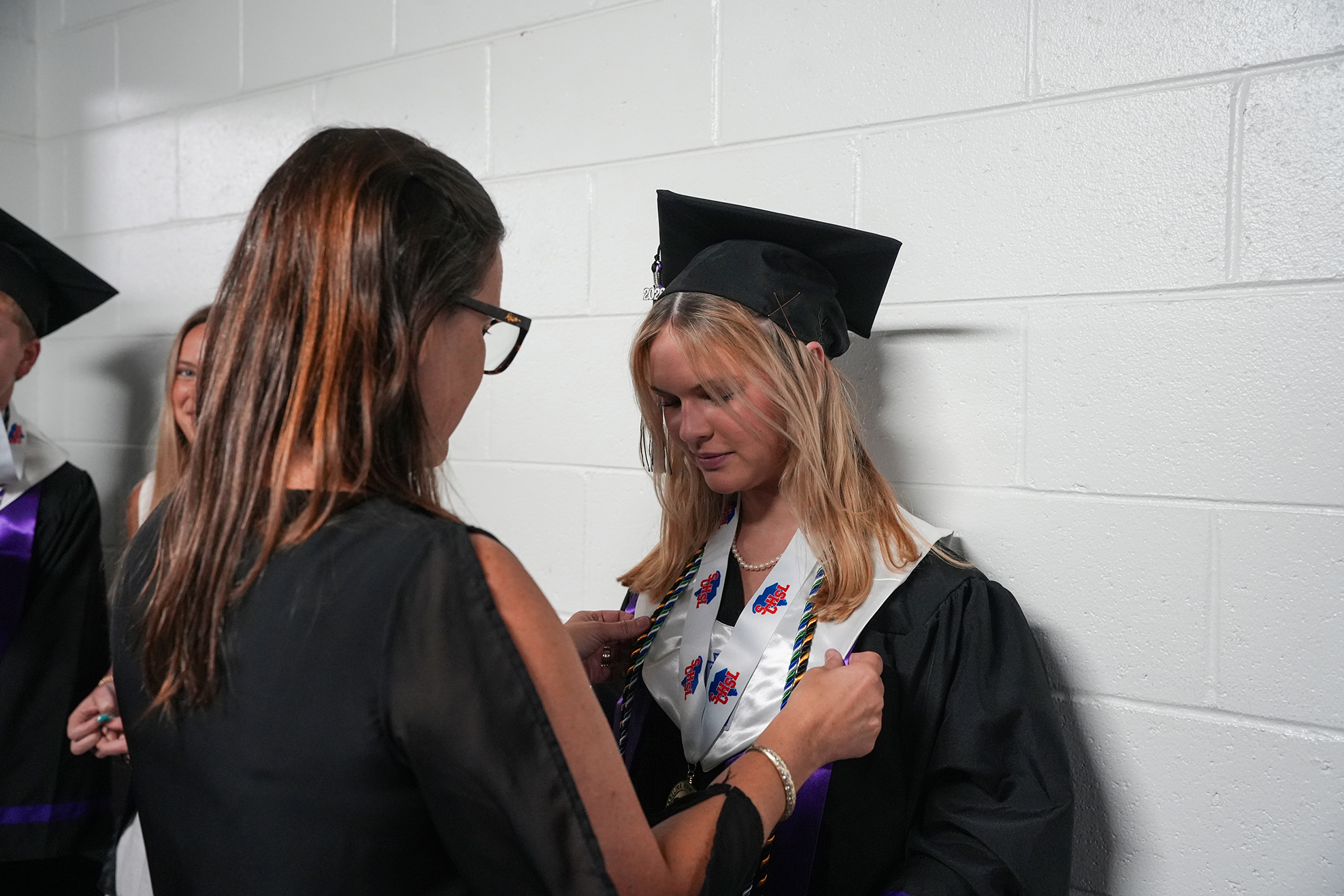 A woman in a black gown adjusts the neck cord of another woman also in a black gown.