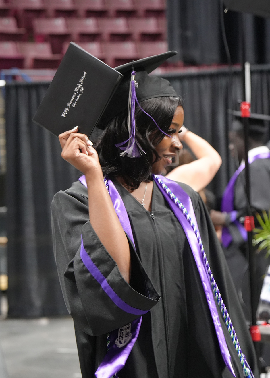 A person in a black and purple gown and cap stands in a room with rows of seats.
