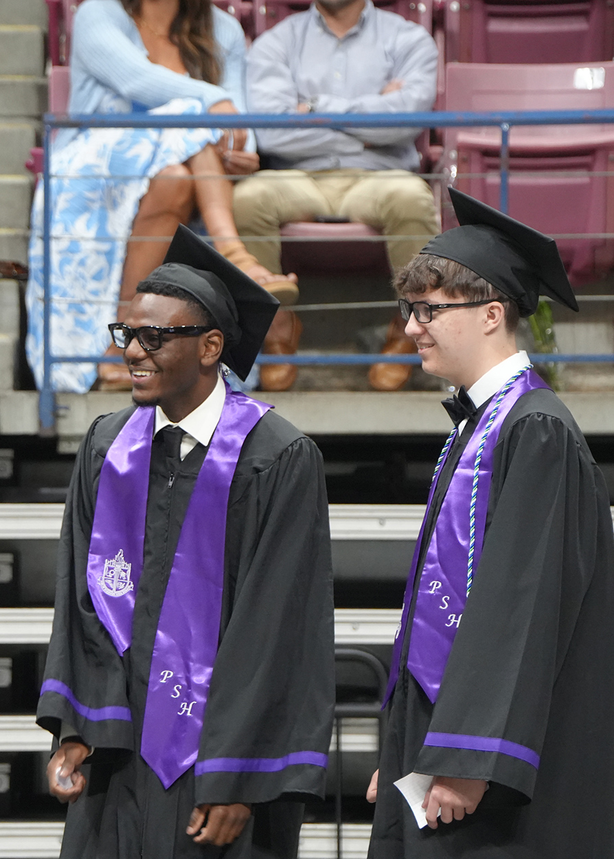 Two young men in graduation robes and caps smile at each other. Behind them, two people sit in bleachers.