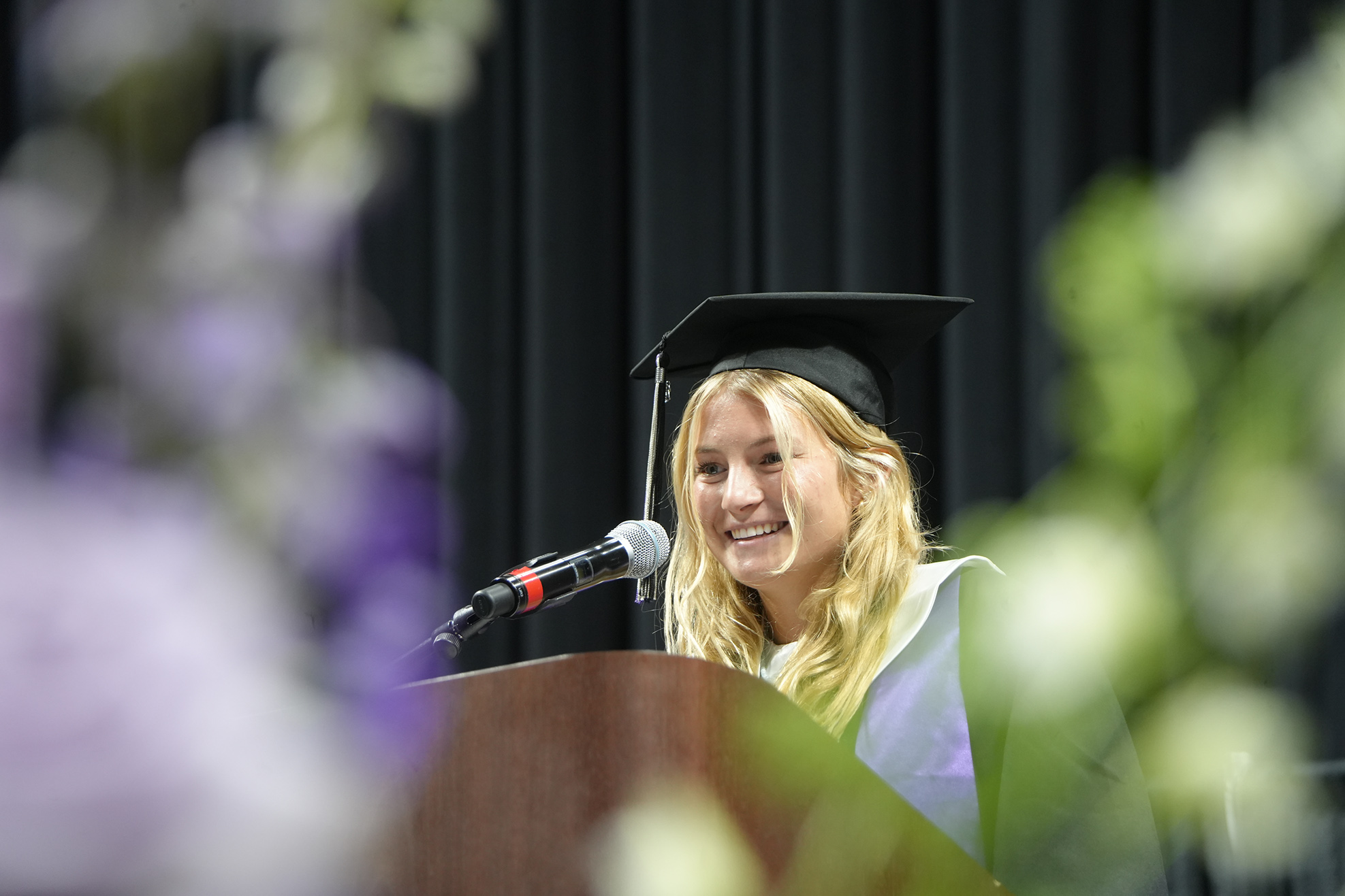 A woman in a graduation gown stands at a podium, speaking into a microphone. Purple flowers are in the foreground.