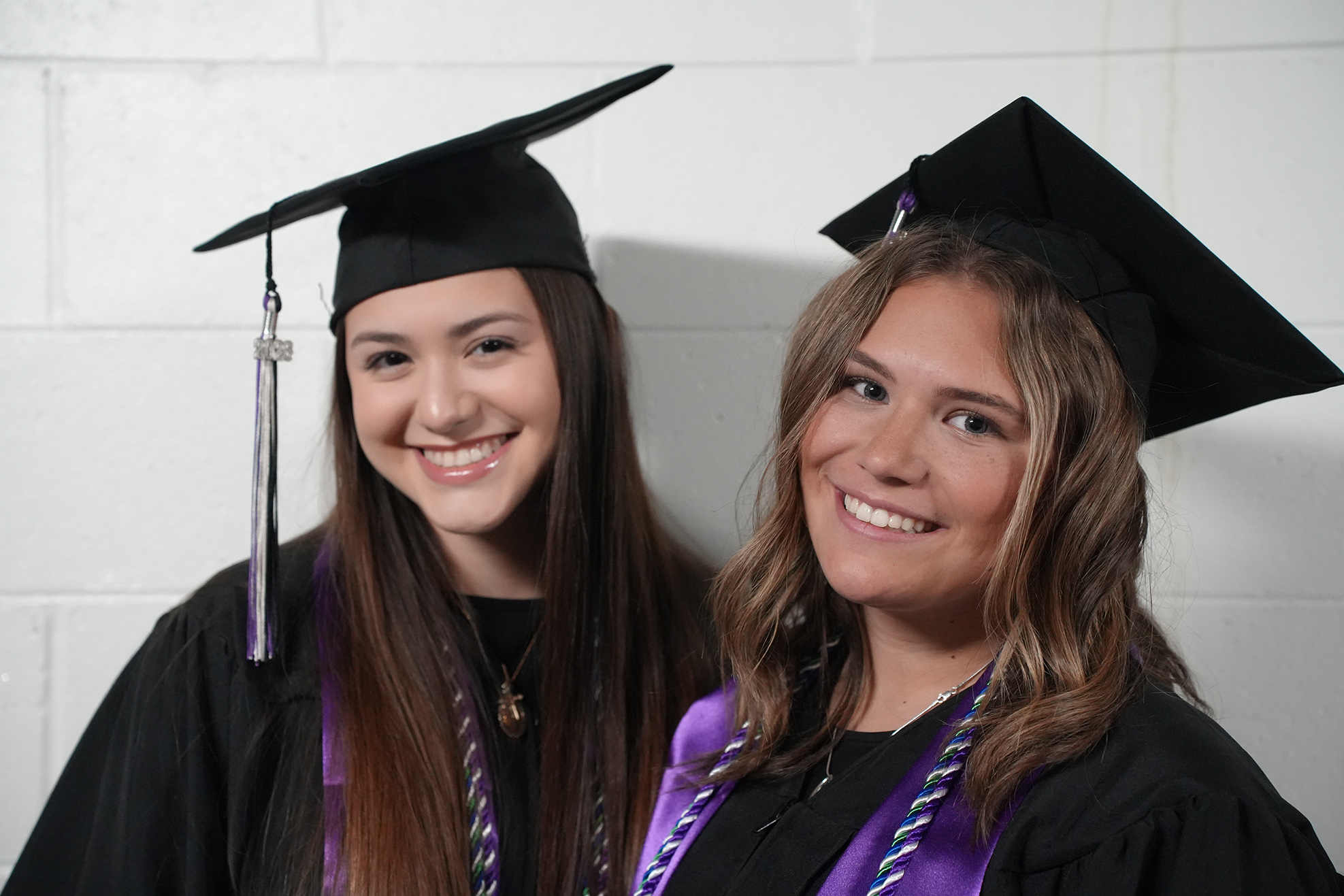 Two women in graduation gowns and caps stand next to each other. Both women smile and wear sashes.