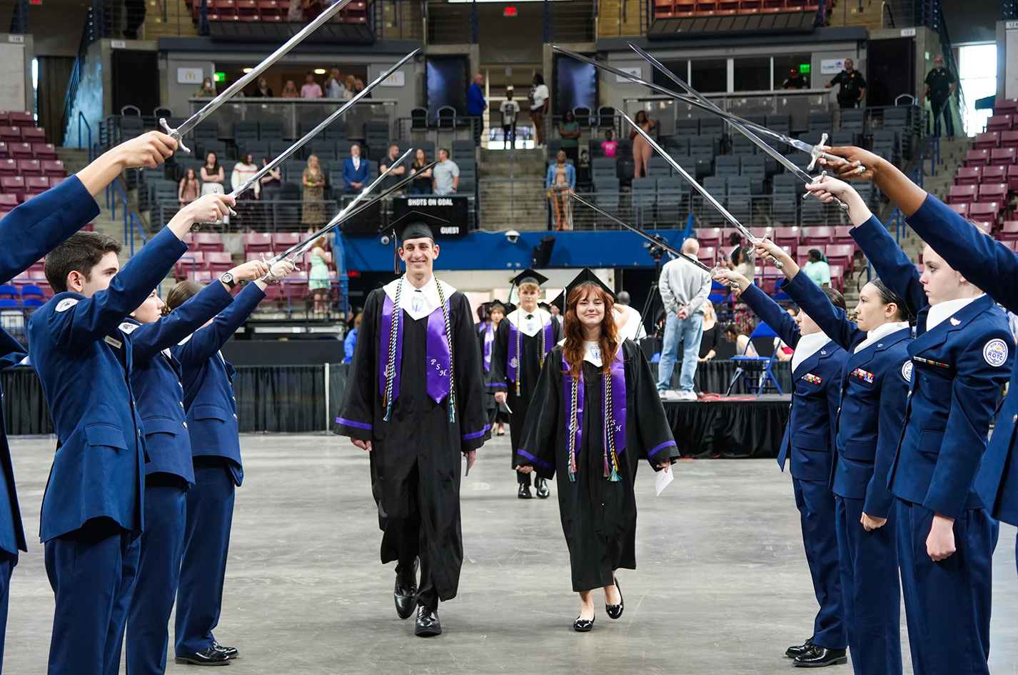 Graduates in blue uniforms walk between two lines of cadets holding swords, in a stadium with empty seats.