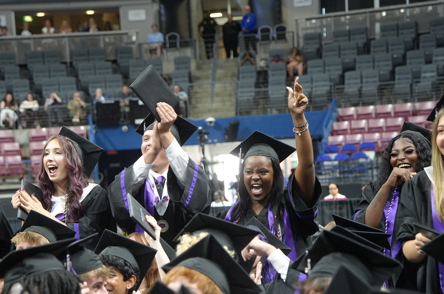 Graduates in caps and gowns, celebrating in a large auditorium with rows of seats. Some are holding up their diplomas.