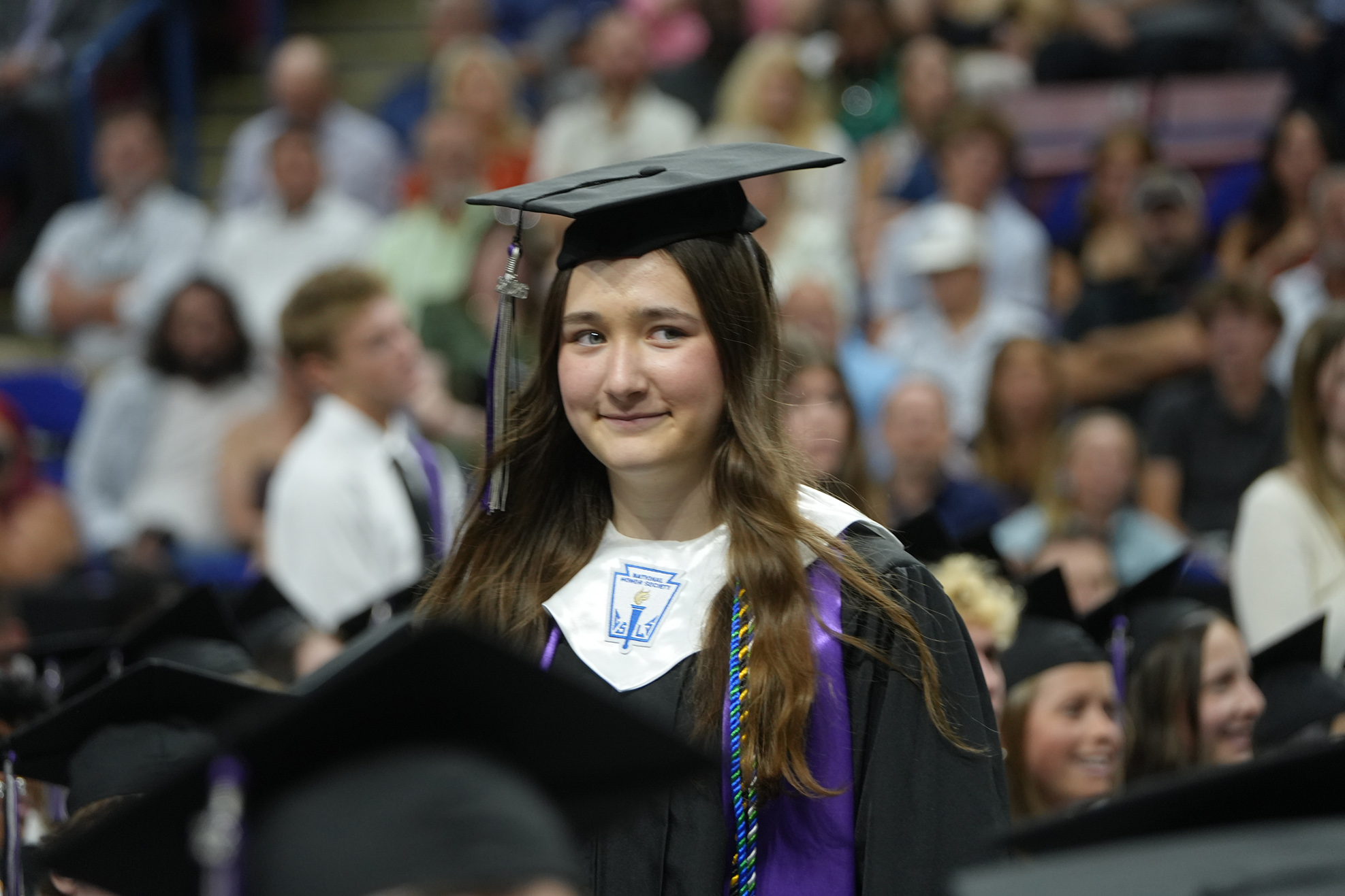 A woman wearing a graduation cap and gown smiles at a graduation ceremony with other graduates.