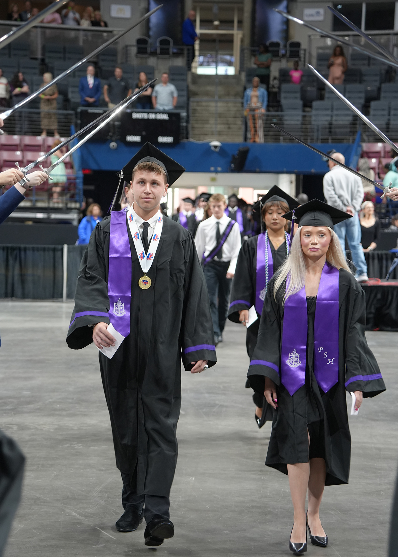 Graduates in black gowns and caps walk in a hall. A man holds a paper and a medal. A woman wears a cap and has blonde hair.