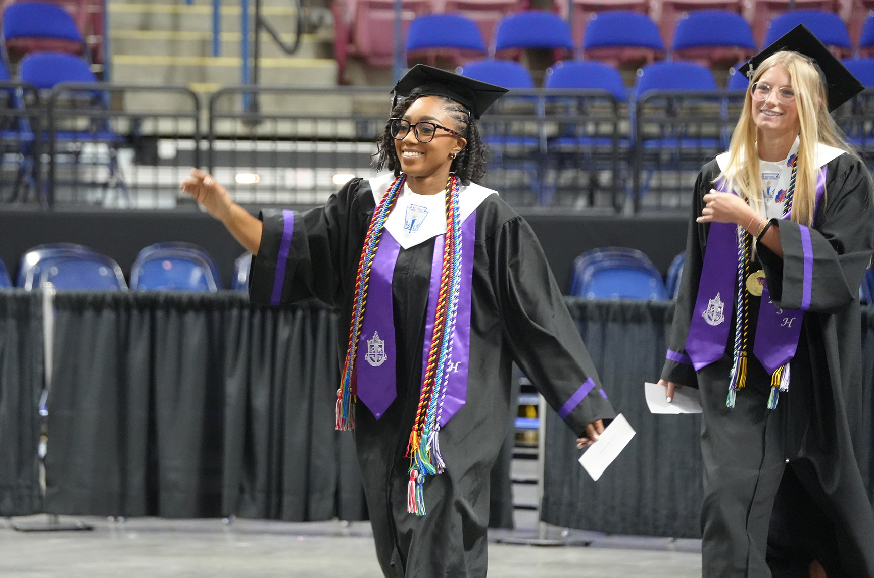 Two women in academic regalia, walking on a stage, one pointing, and the other holding a paper.