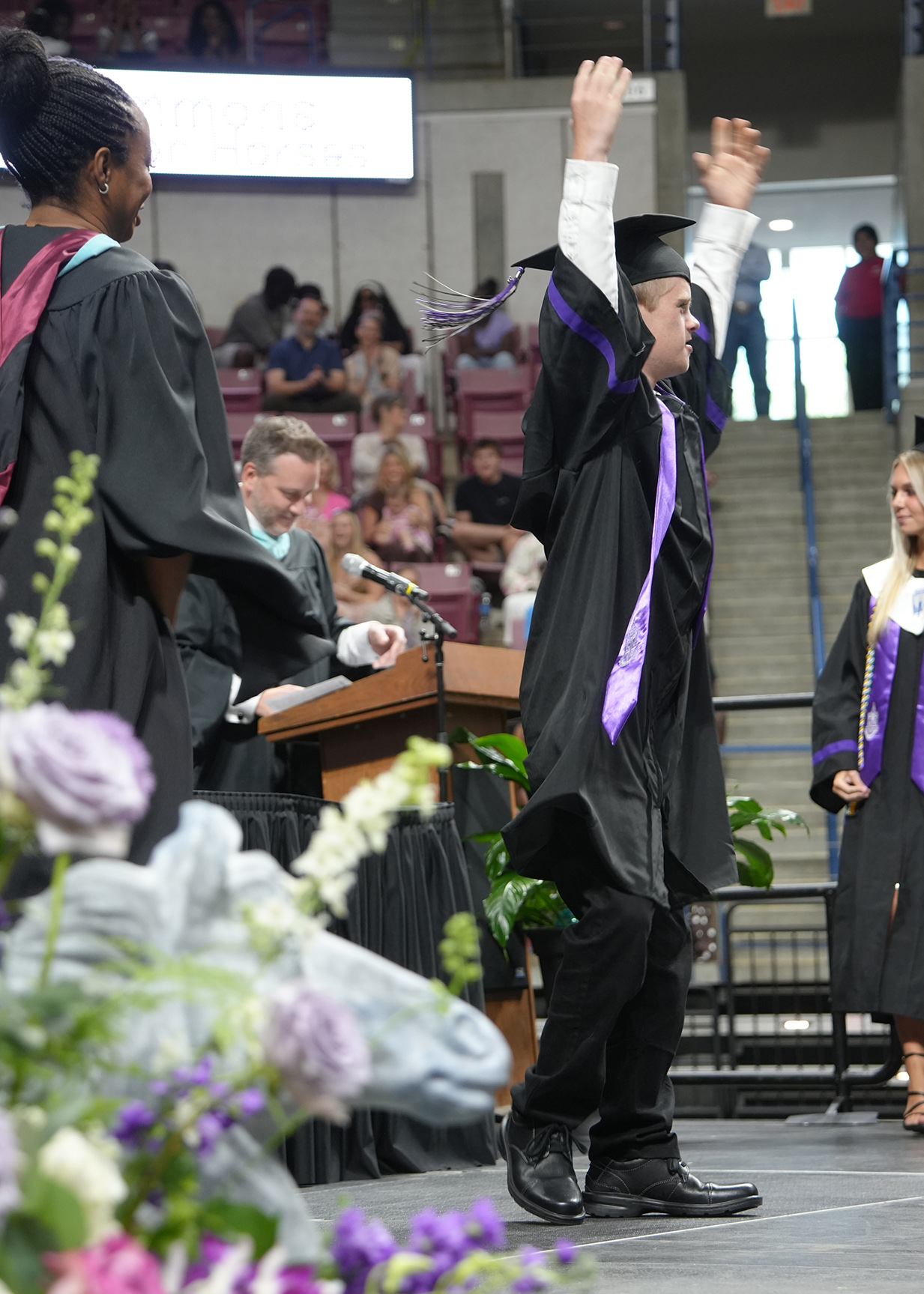 A person in a cap and gown celebrates on stage at a graduation ceremony with flowers.