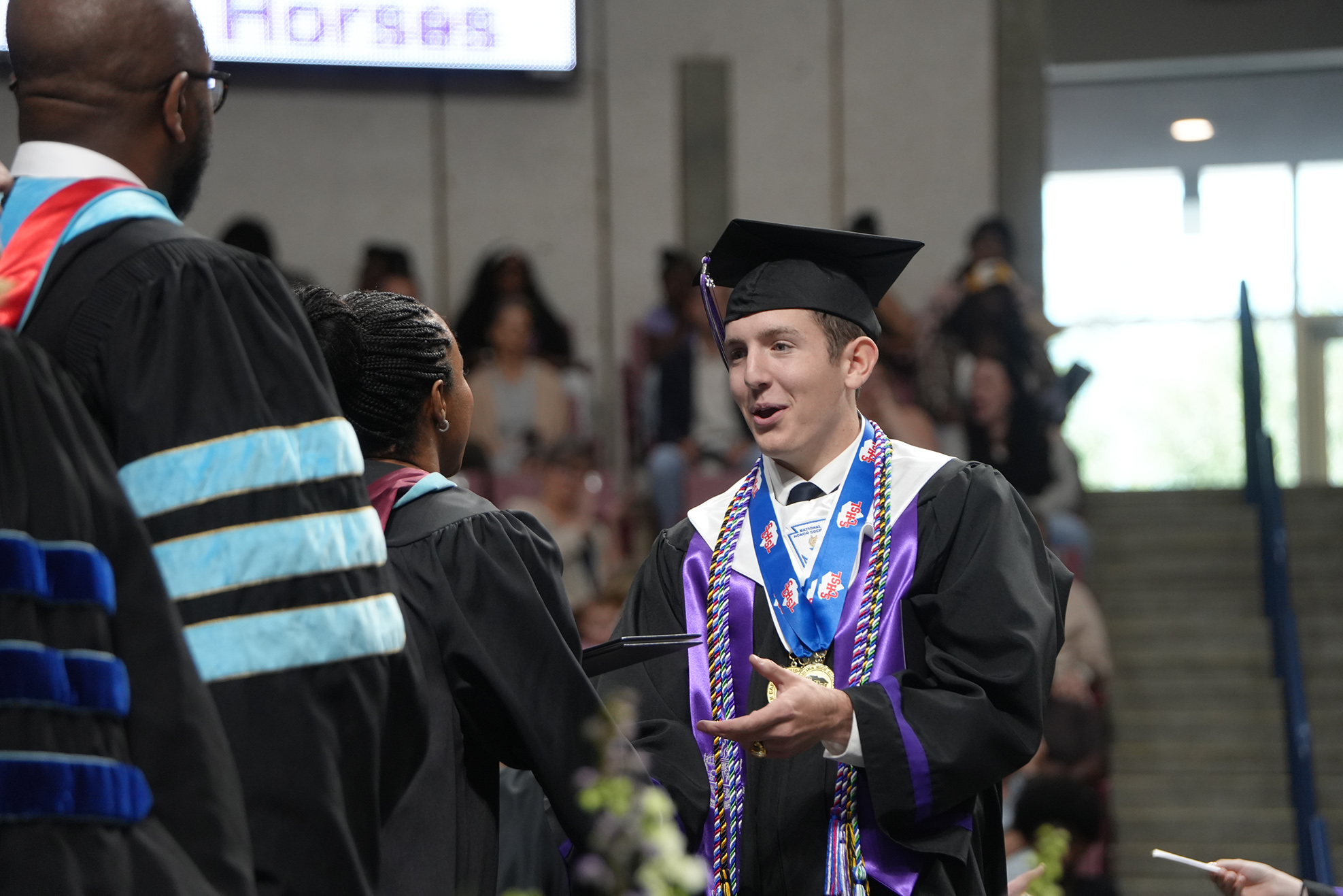 A man in a black gown and cap stands, smiling, with a medal around his neck. Behind him, others in academic robes stand and converse.