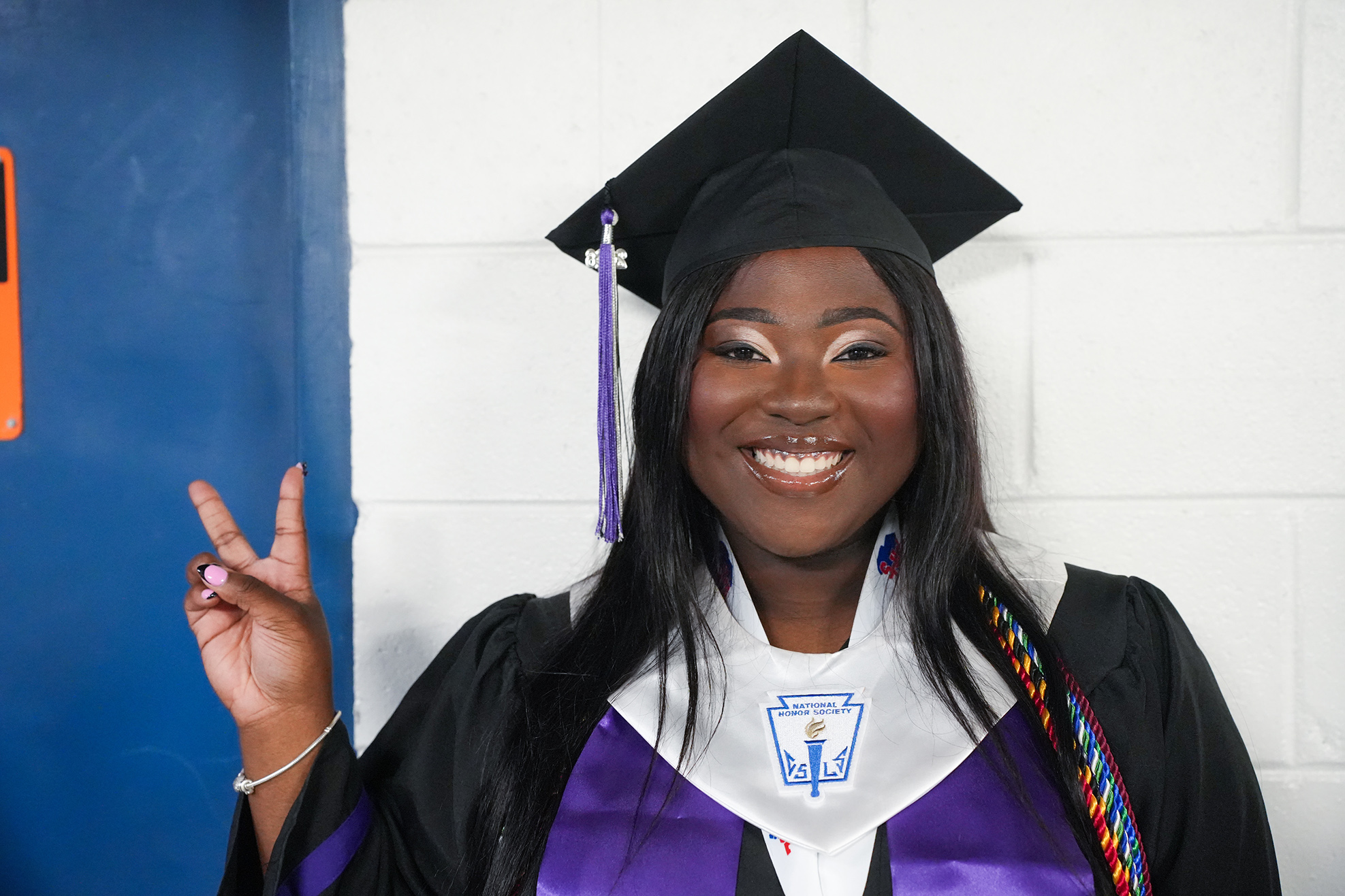 A woman in a graduation gown with a tassel smiles and makes a peace sign with one hand.