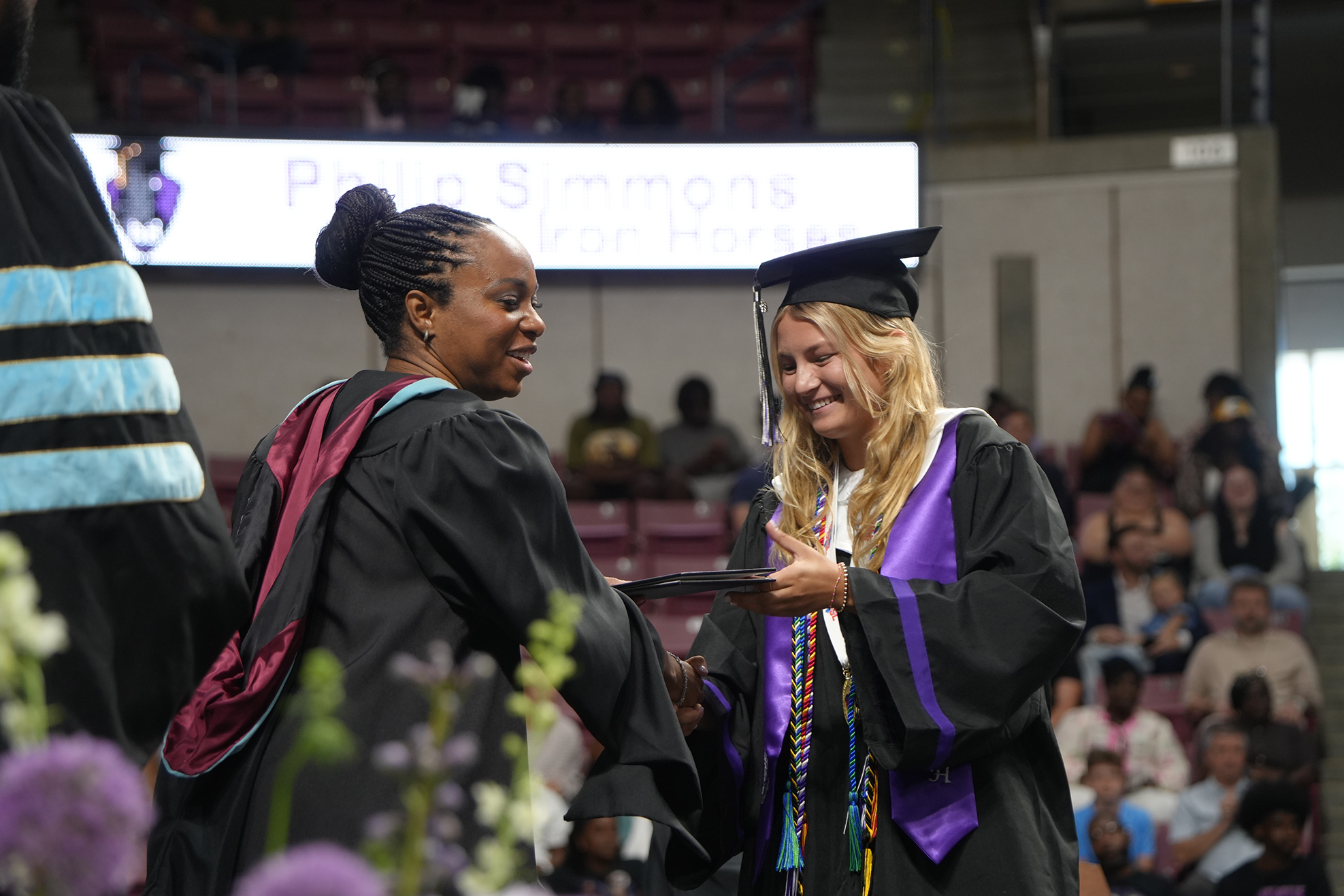 Two women in academic robes, one with a tassel, stand before a sign. They smile and exchange an item. Behind them, an audience sits in bleachers.