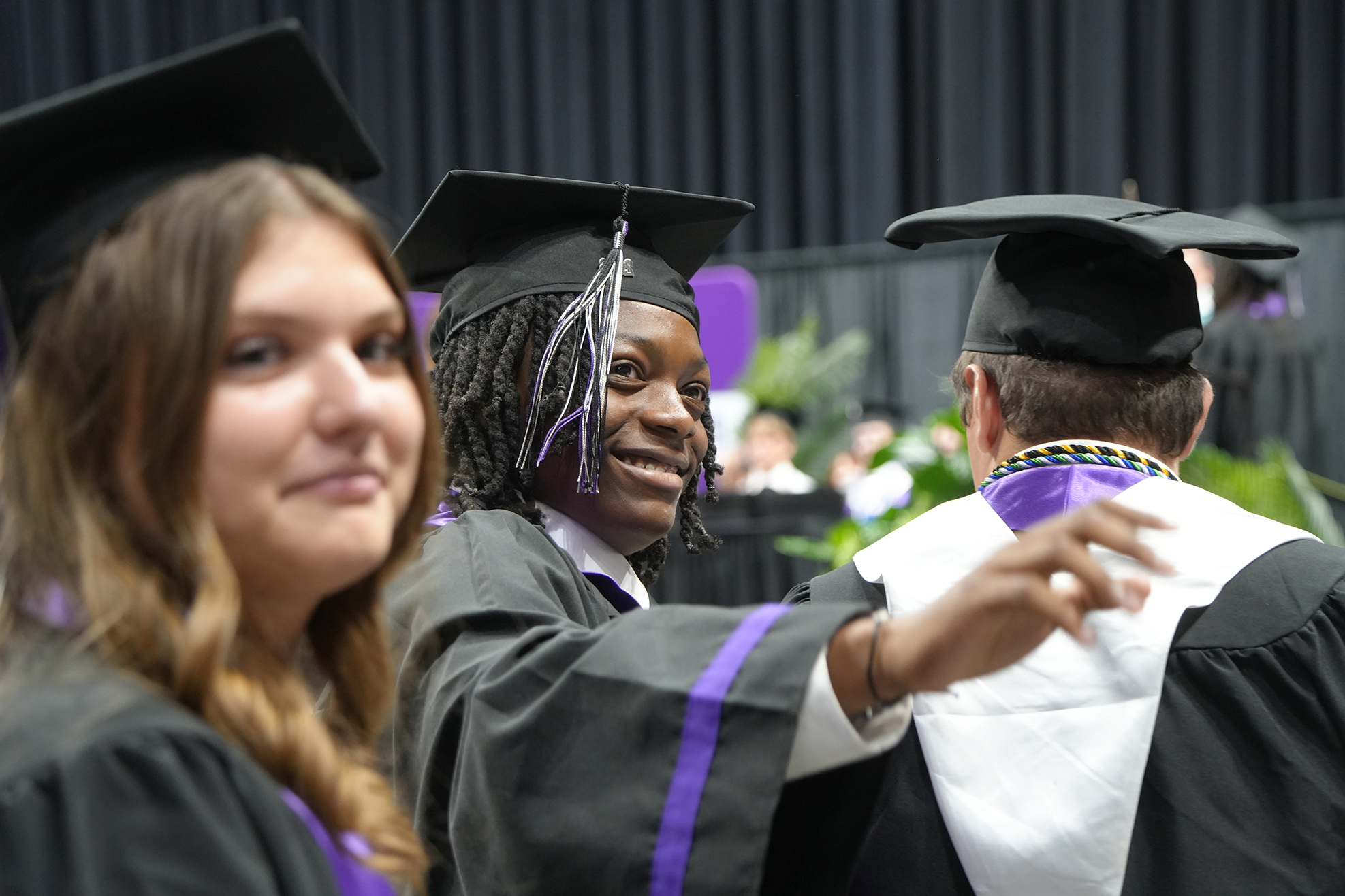 Three individuals wearing black graduation gowns with purple trims and mortarboards. They are at a graduation ceremony.