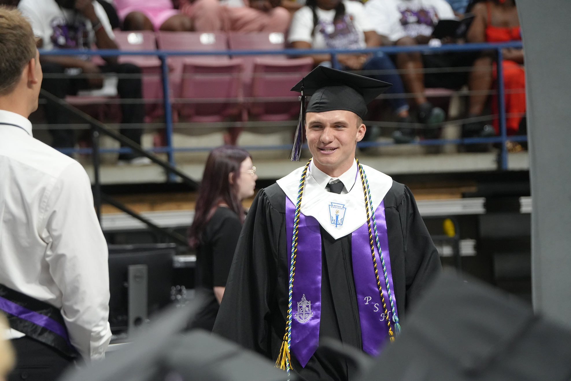 A graduate in a cap and gown walks down the aisle of an auditorium, with audience members seated in bleachers.