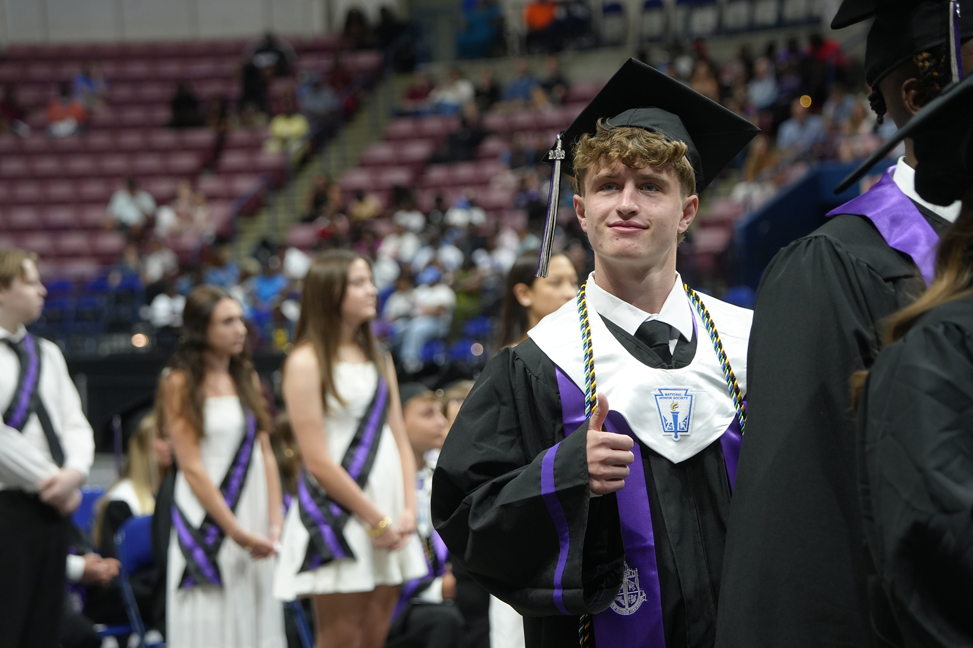 Graduates in black gowns, one giving a thumbs-up, stand in a stadium with seated audience.