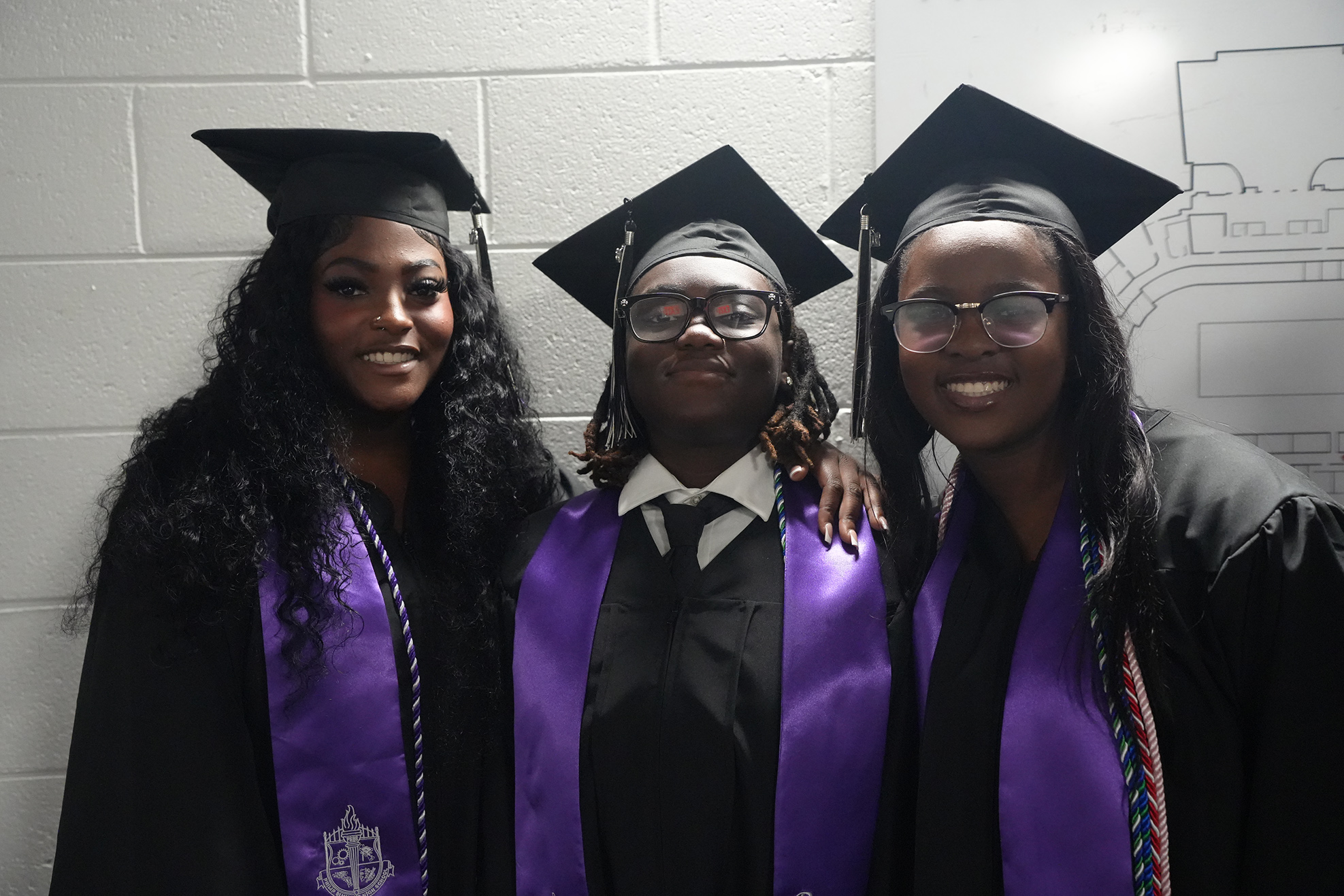 Three women in graduation attire, one in a purple gown, pose together against a white wall.