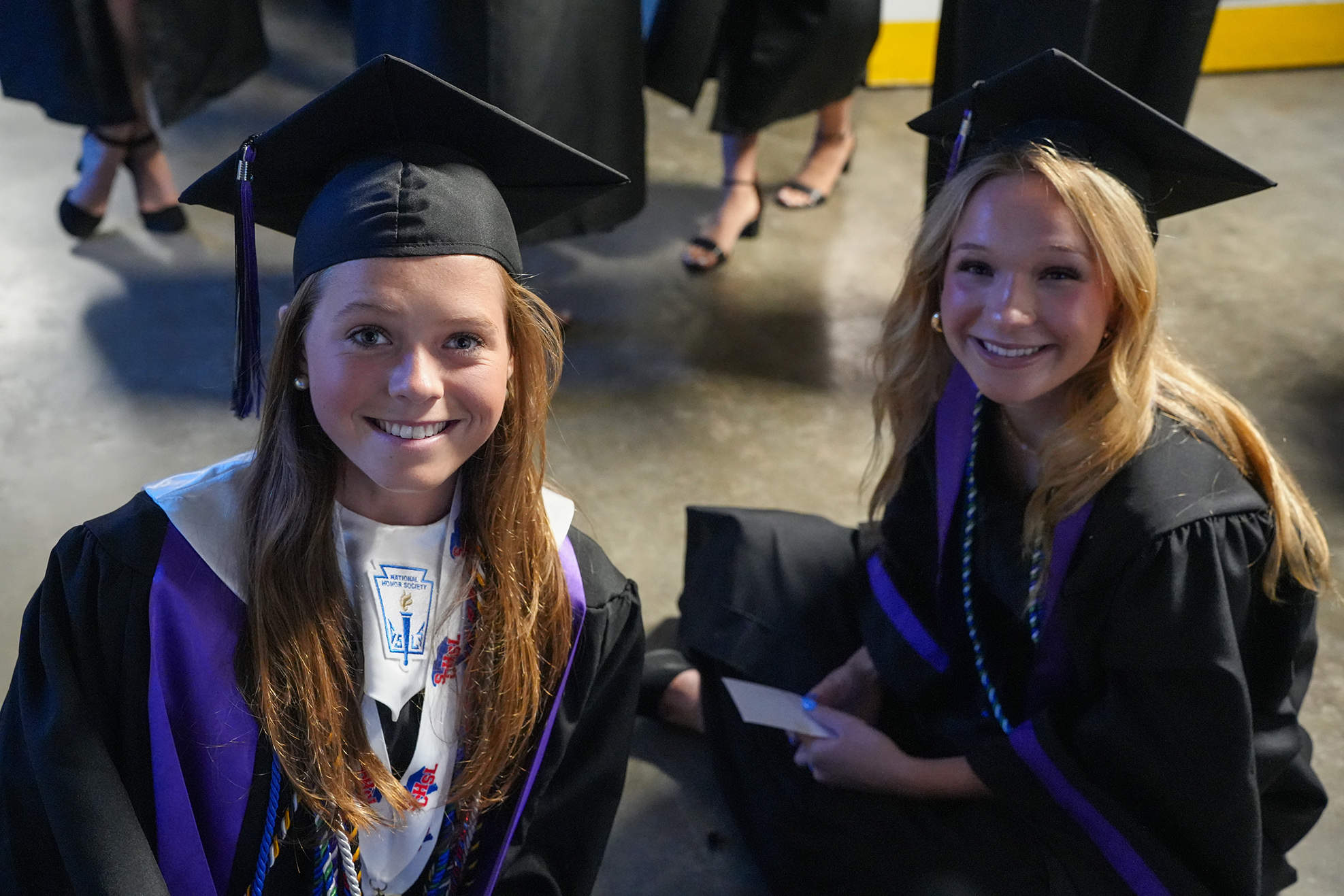 Two young women in black graduation gowns, one sitting and one standing, smile for the camera.