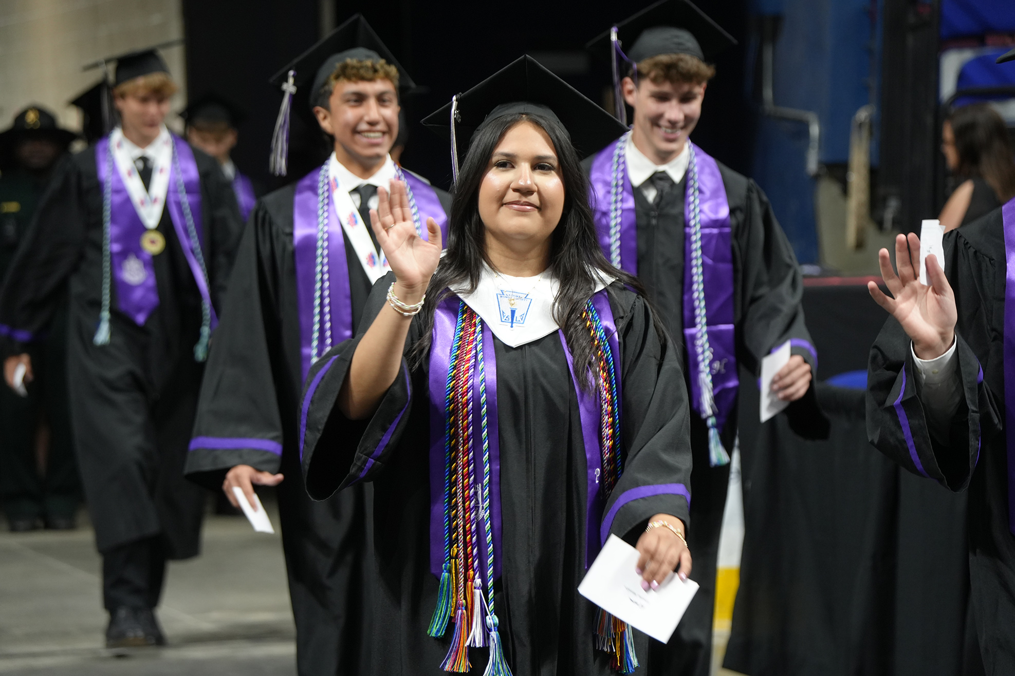 A woman in a graduation gown waves as she walks past others wearing caps and gowns.
