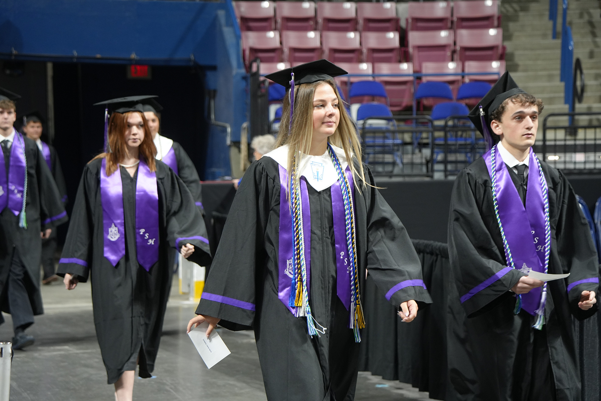 People in black and purple graduation gowns and caps walk in a line in an empty auditorium.
