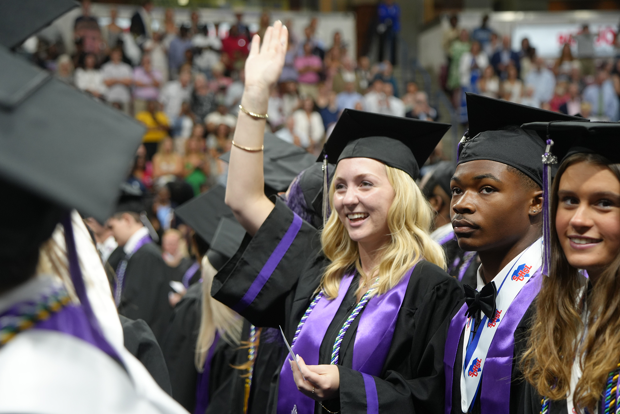 Graduates in gowns and caps, one waving, smile at the camera; a large audience in the background.