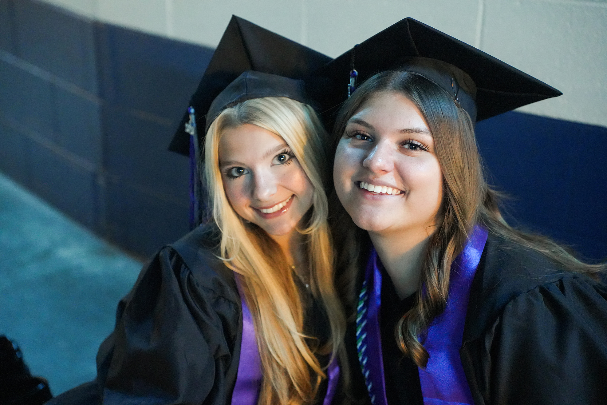 Two women in caps and gowns smile at the camera, seated against a wall with a blue pattern.