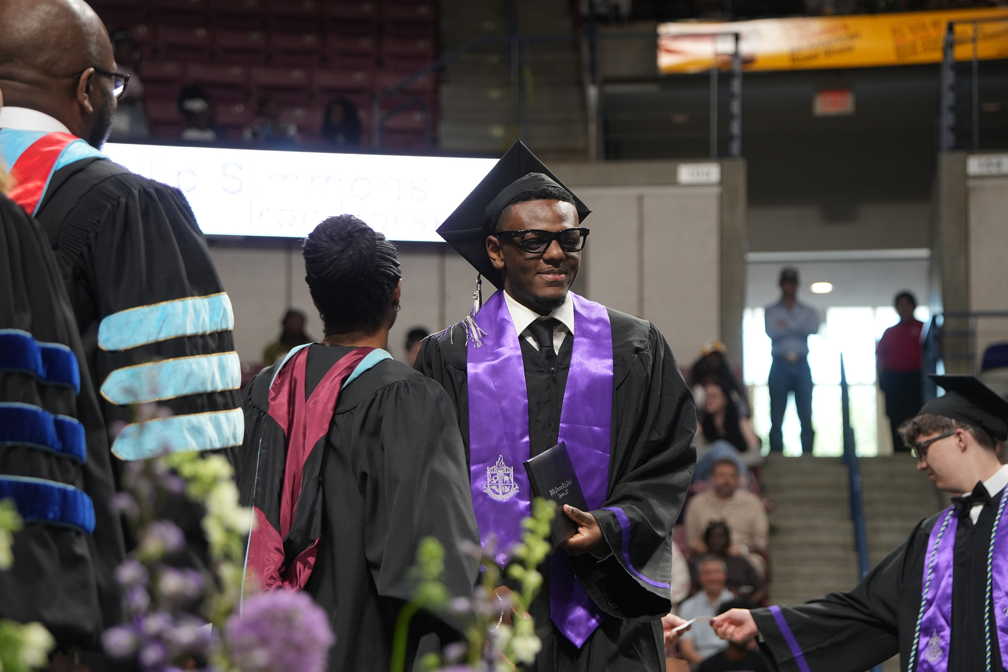 Graduates in black gowns with stoles of various colors stand in a stadium, some with caps and glasses.