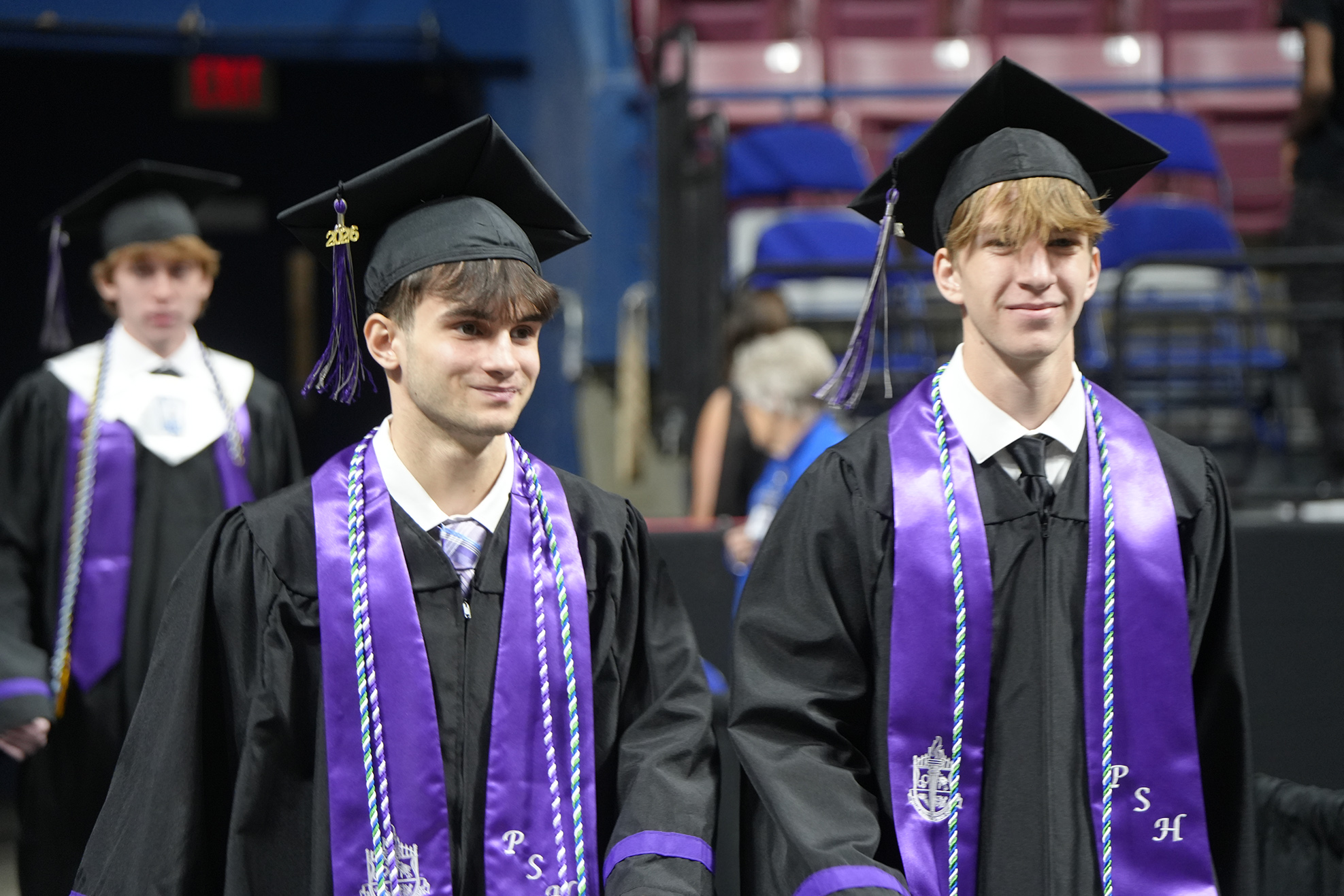 Two men in black graduation gowns and hats walk in a stadium; blurred background shows empty blue chairs and a crowd.