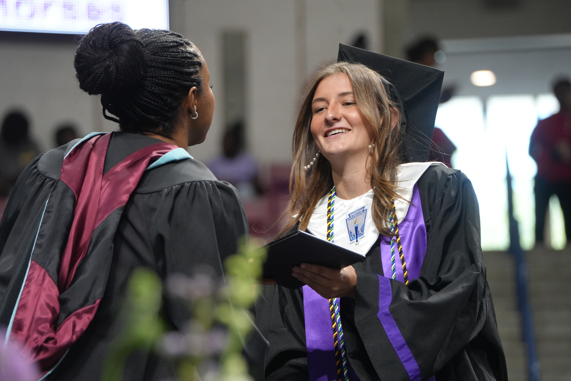 Two women in academic regalia converse at a graduation ceremony. One holds a book. Behind them, blurred people are present.
