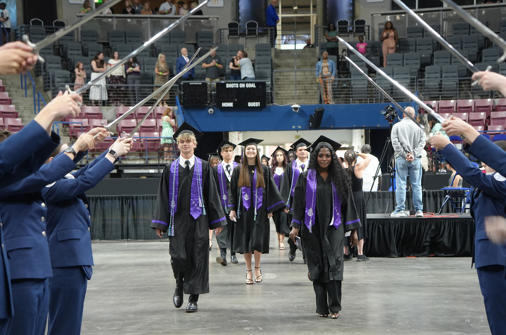 A group of people in blue uniforms and caps walks on a stage, holding swords, with spectators in the background.