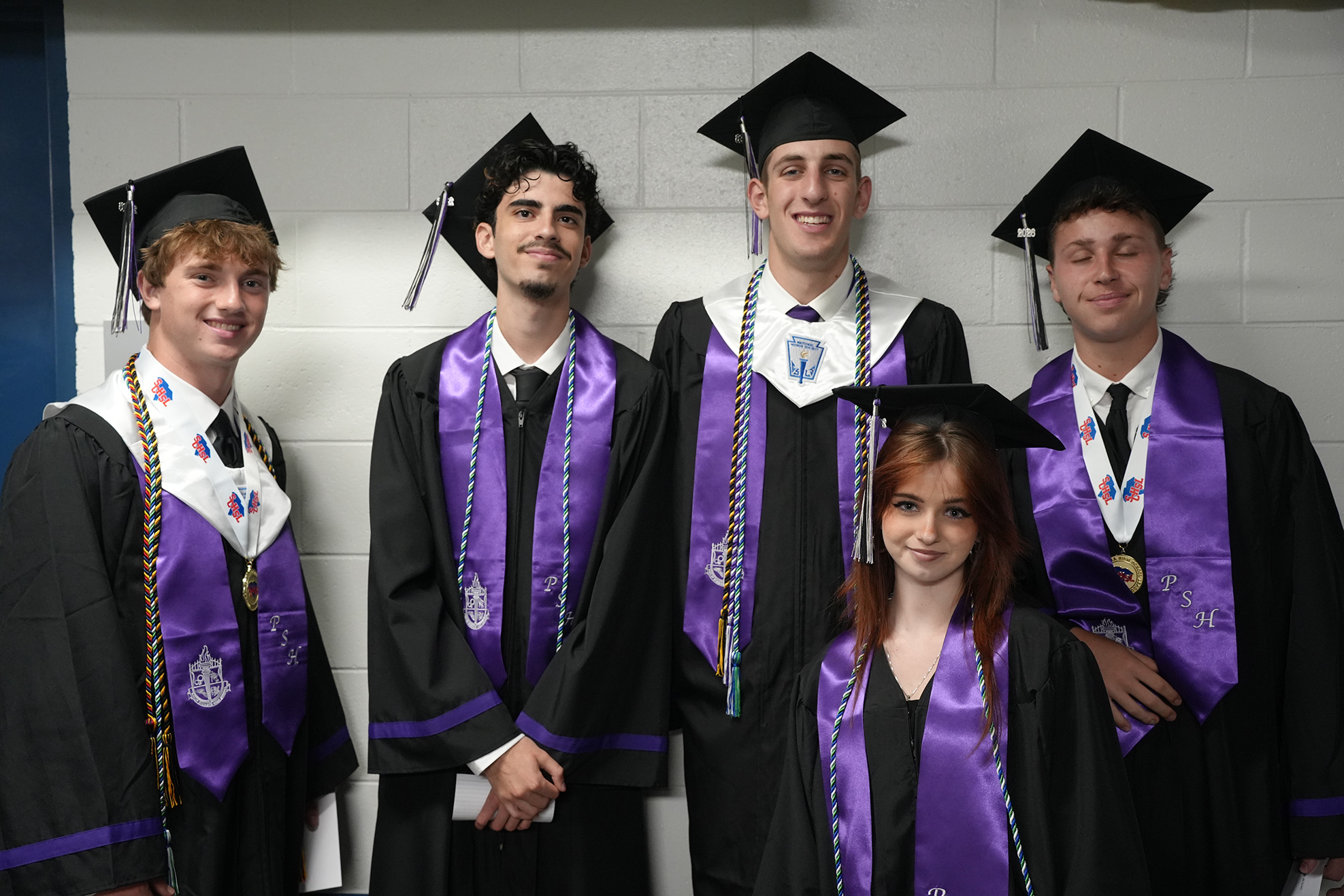 Five individuals in black graduation gowns and hats stand close together, with three wearing purple sashes.