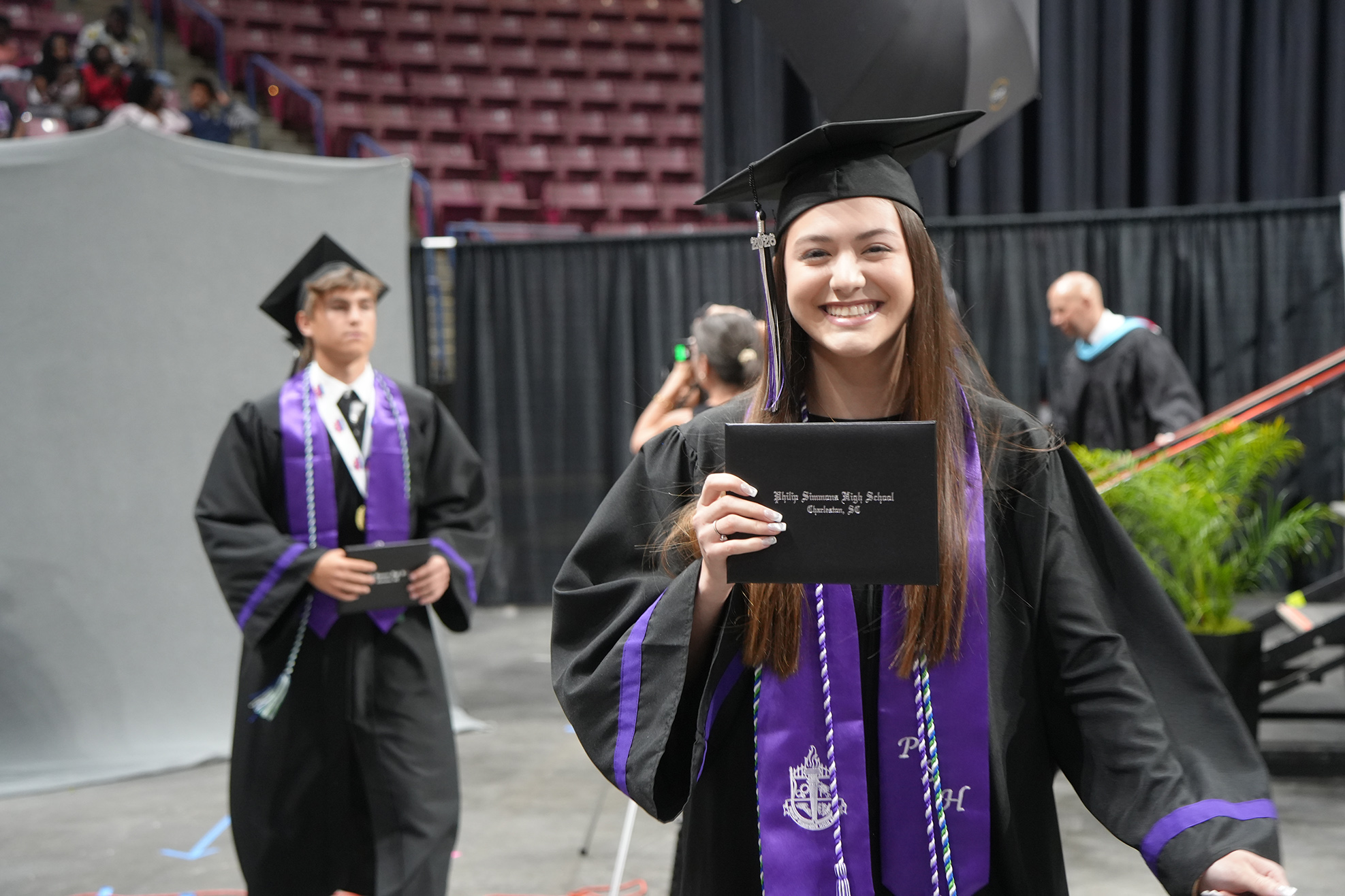 A smiling graduate holds her diploma, wearing a cap and gown, with another graduate in the background.