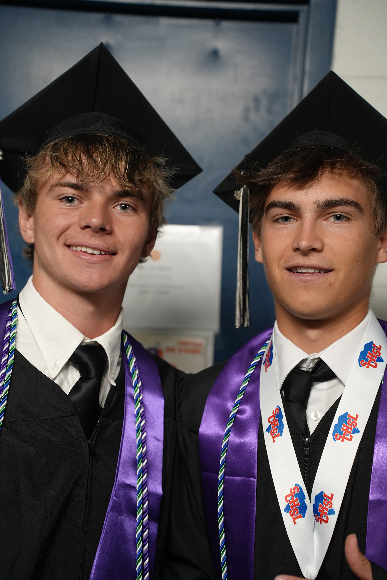 Two young men in academic regalia, smiling, one with a tassel and the other with a sash.