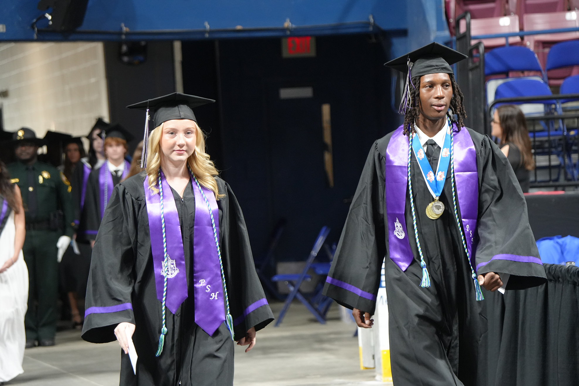 A woman and a man in academic regalia walk forward, smiling. They wear purple sashes and medals. Behind them, others in robes walk on a stage.