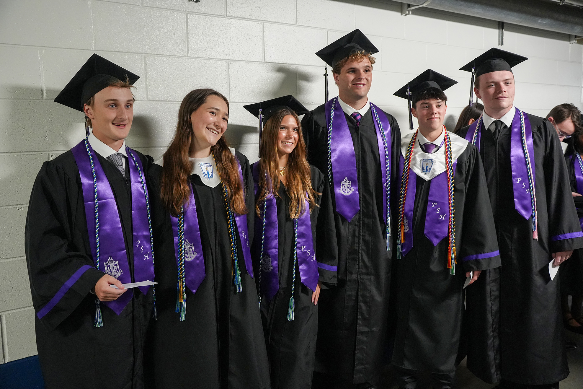 Six young adults in academic regalia, smiling and standing together against a white wall.