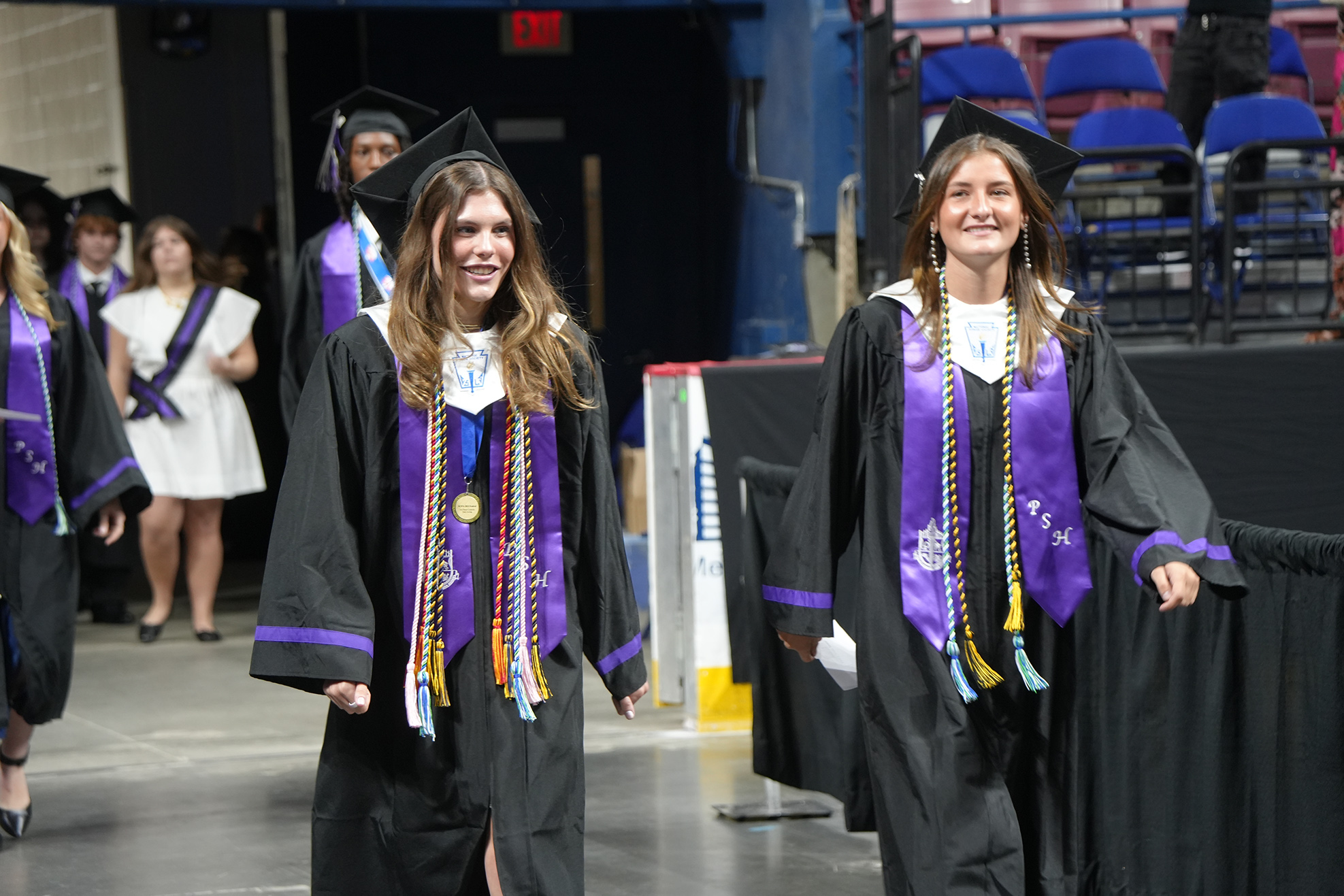 Two women in black gowns and mortarboard hats walk in a line with other graduates at an indoor event.
