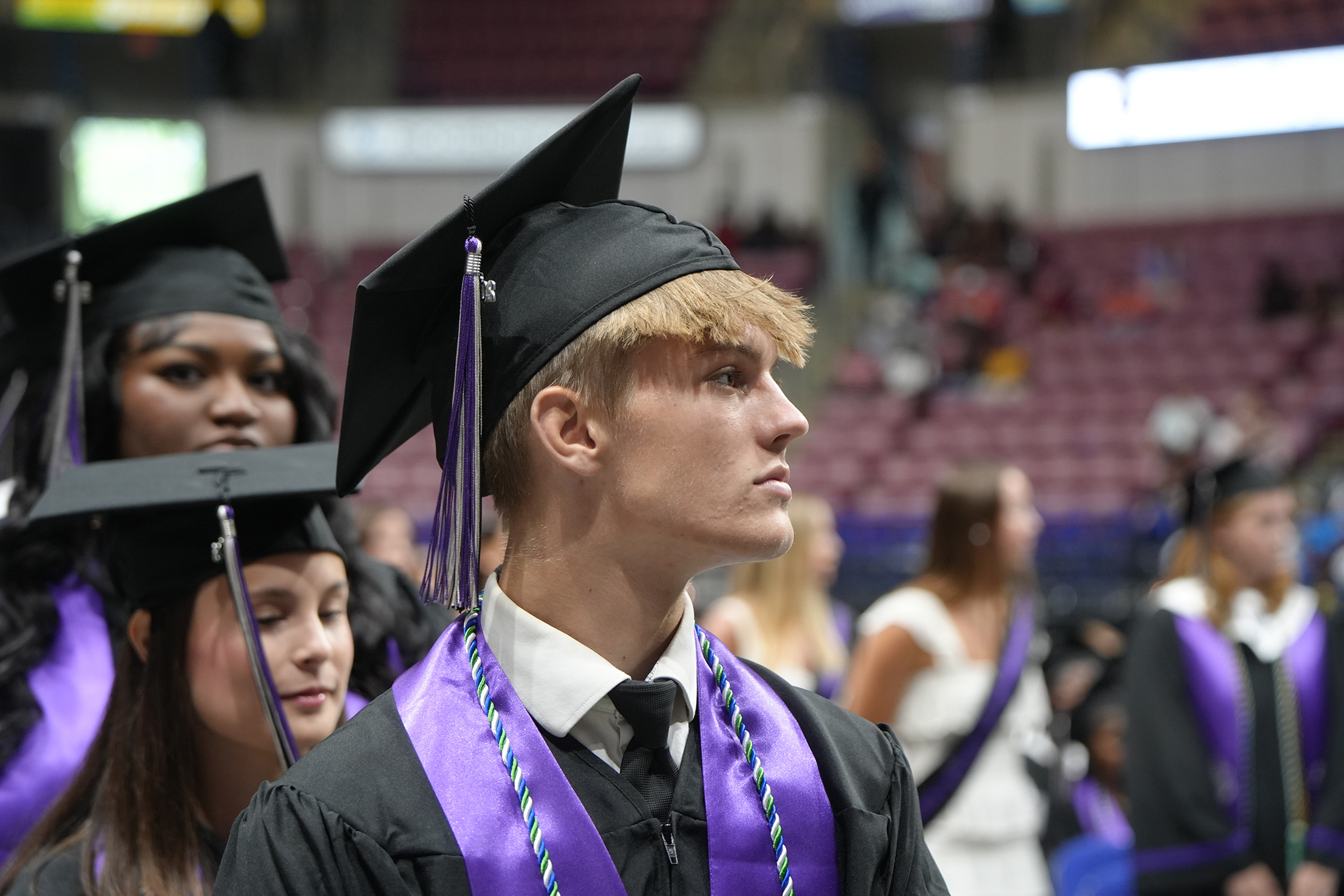 Graduates in caps and gowns stand in a stadium, with rows of seats in the background.