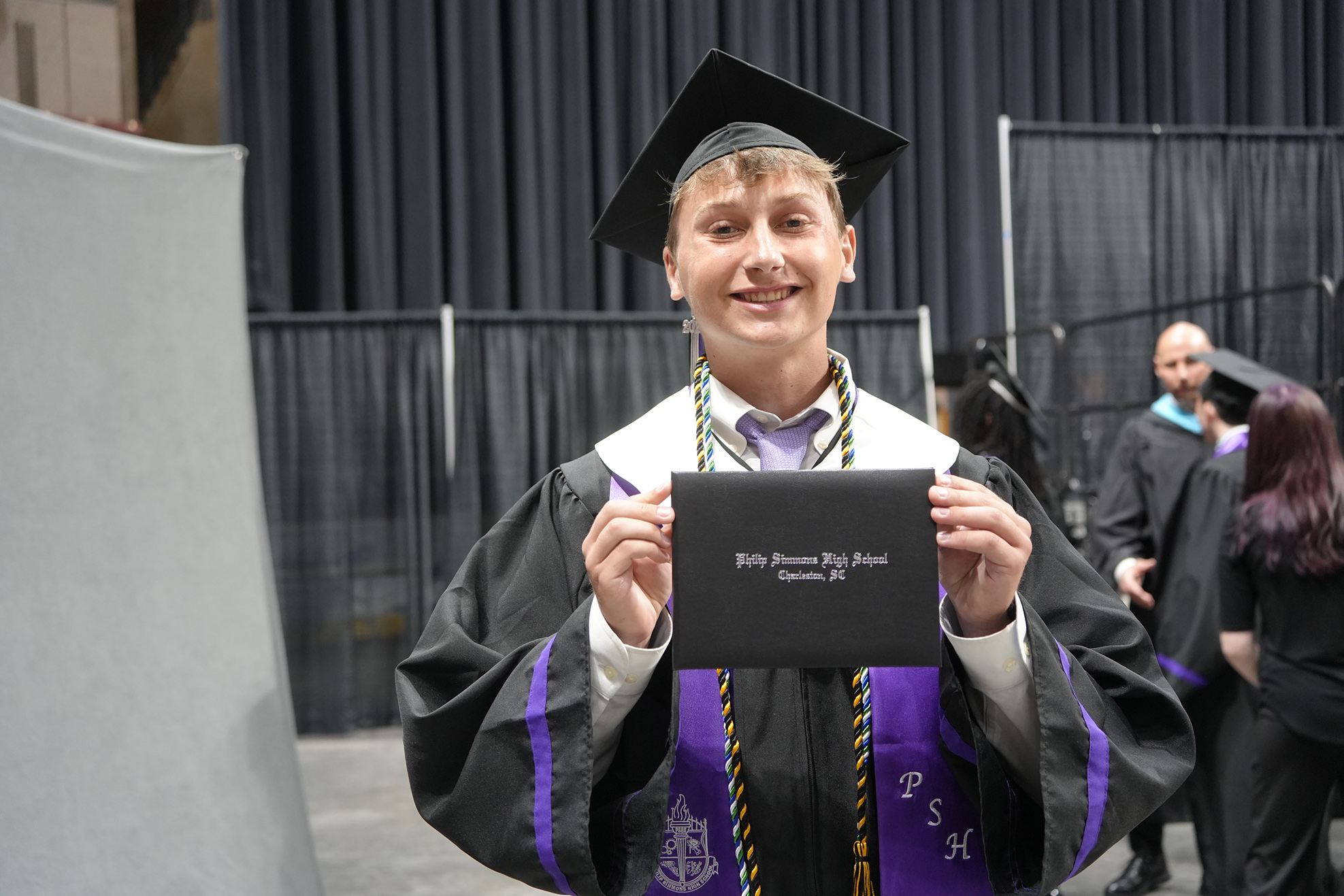 A young person in a graduation gown and cap holds a diploma in both hands, smiling.
