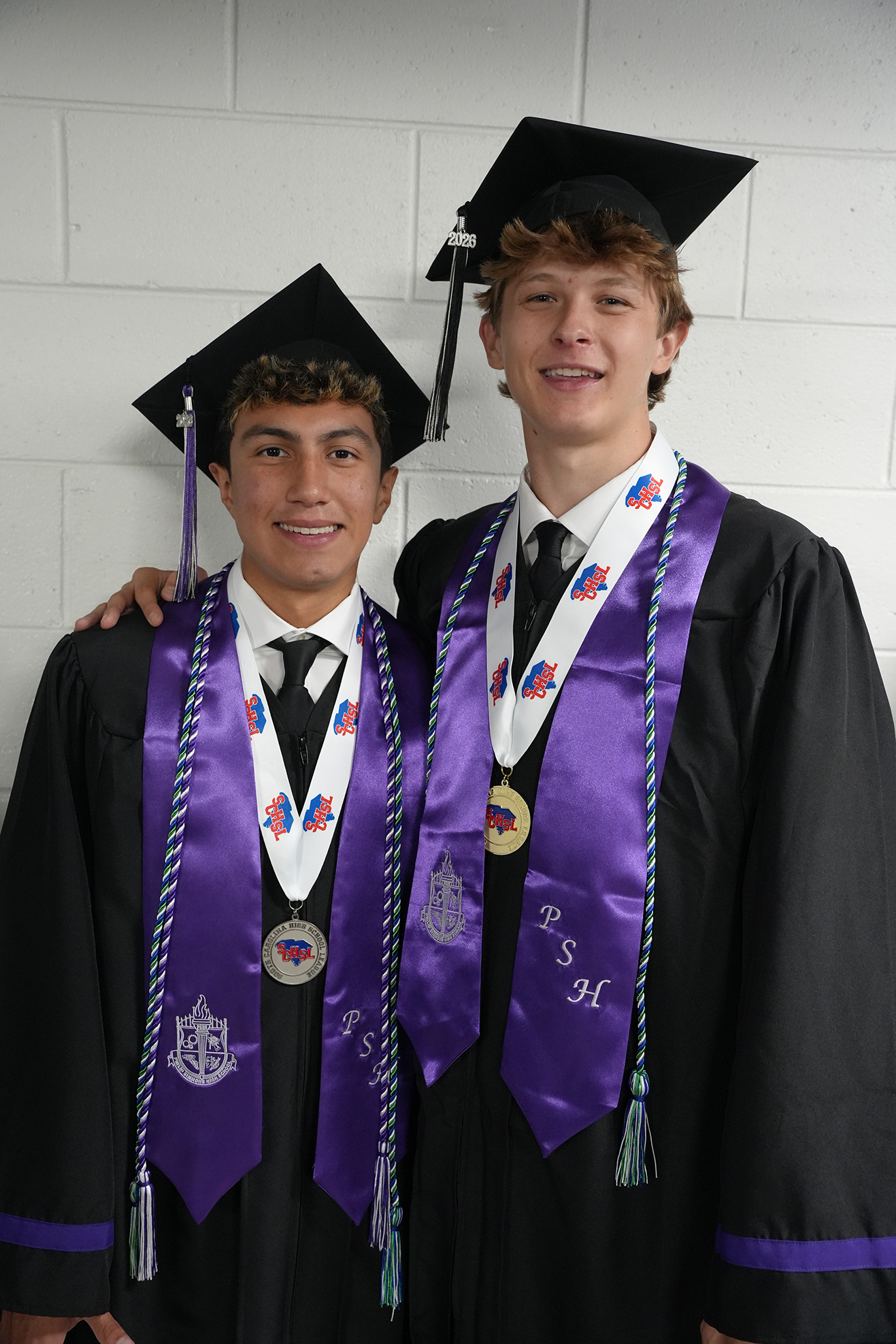 Two men in graduation caps and gowns stand side by side, one with his arm around the other's shoulder.
