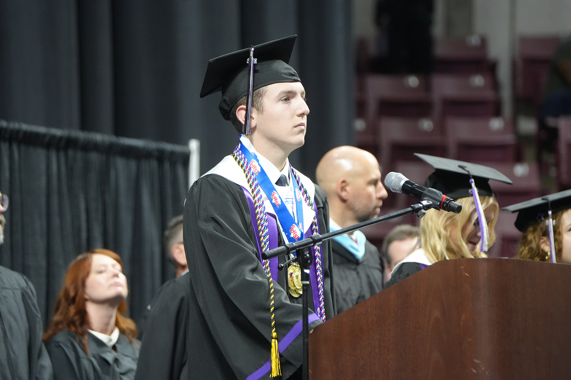 A man in a cap and gown speaks at a podium during an outdoor graduation ceremony with rows of seats behind him.