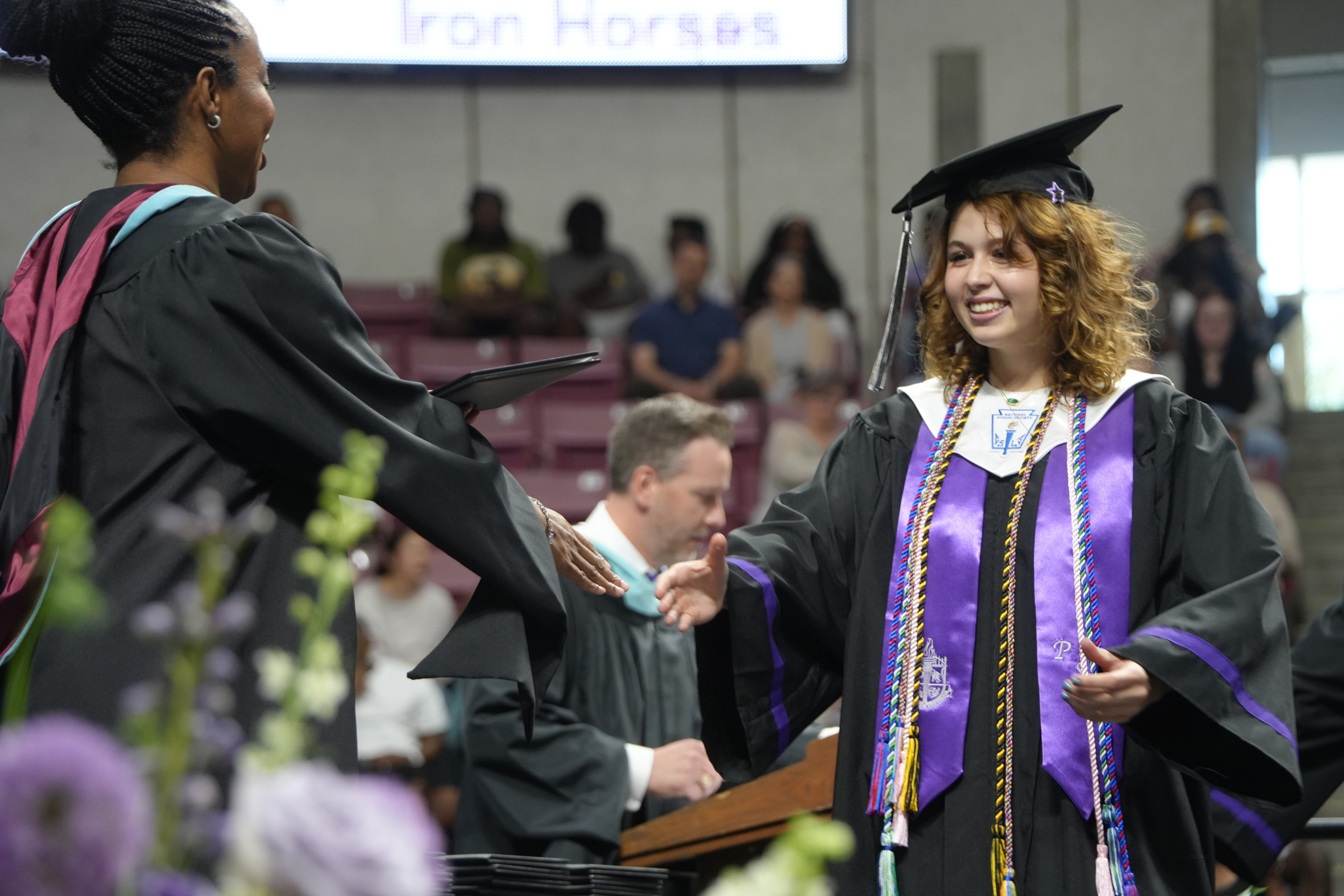 Two women in academic regalia shaking hands at a graduation ceremony. The background shows a large audience.
