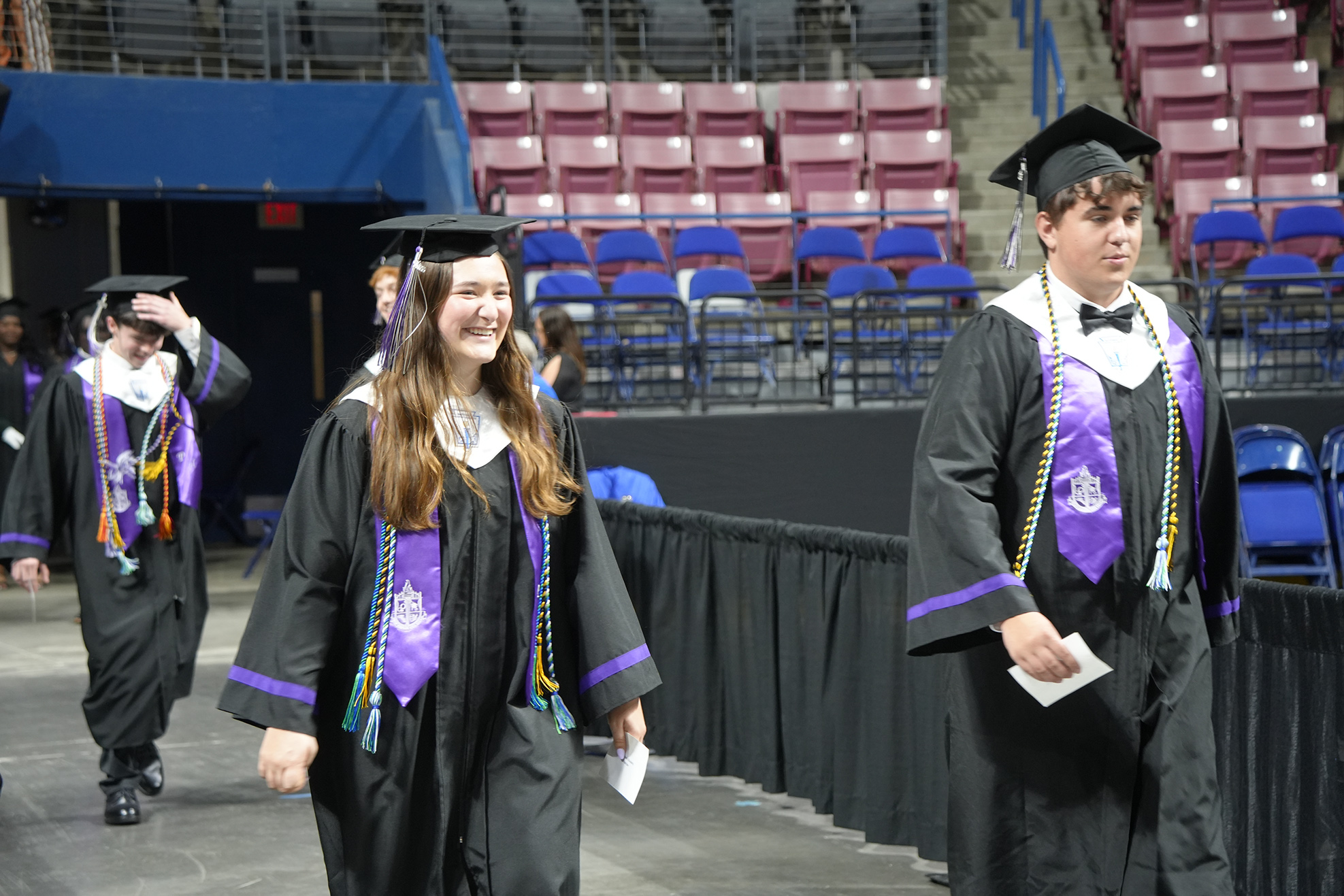Graduates in black gowns and caps walk on a stage with empty red seats in the background.