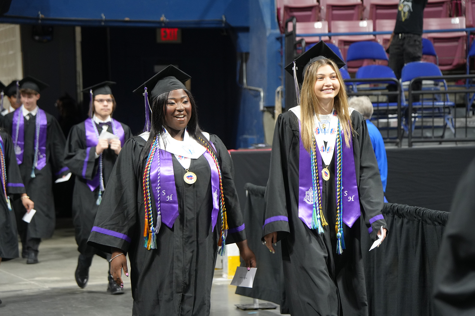 Several people wearing caps and gowns walk through a venue. Some are smiling and appear to be graduating.