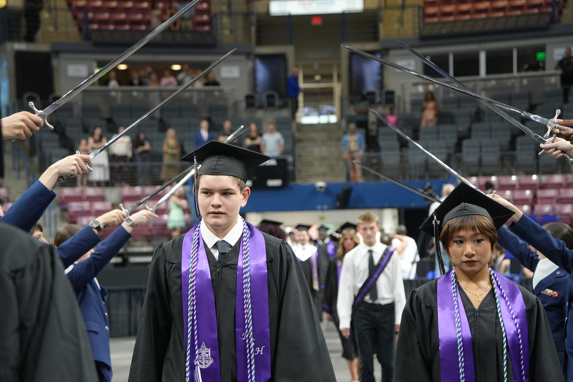 Graduates in caps and gowns holding swords walk across the stage in a stadium with audience members.