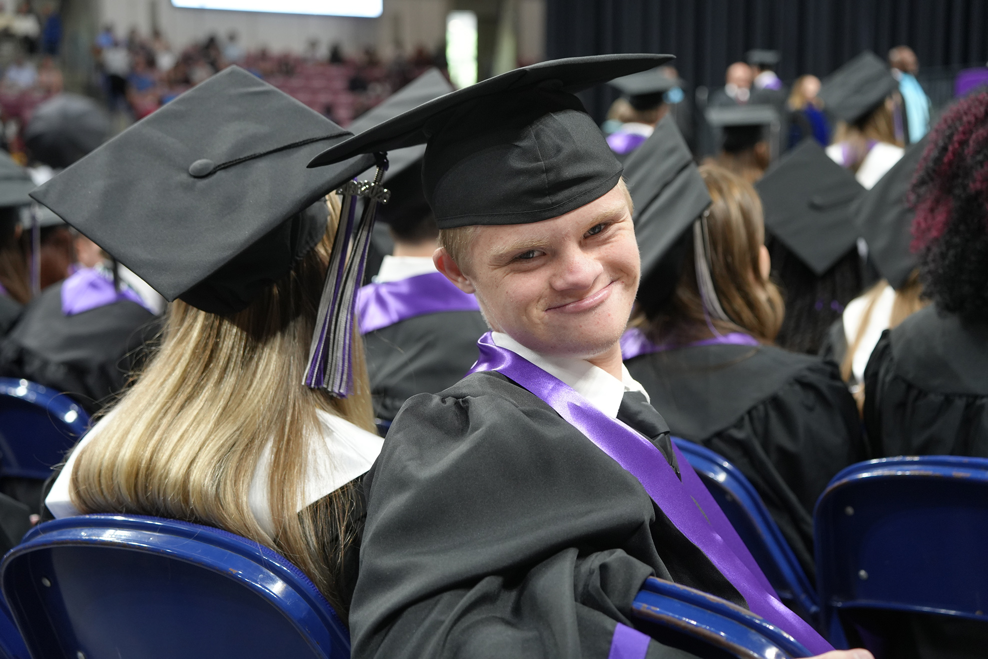 A person in a graduation gown sits in a blue chair with a smiling face.