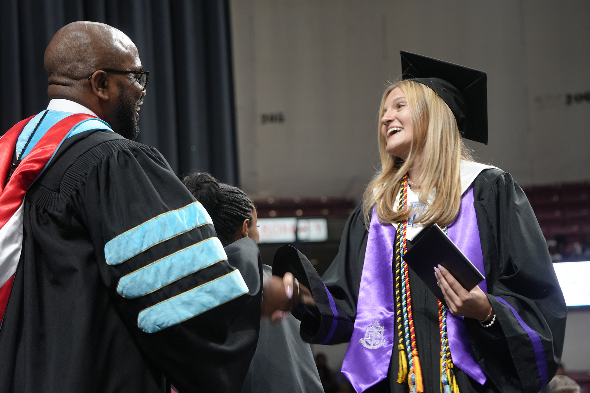 A graduate wearing a cap and gown, smiling, shaking hands with a faculty member in a similar outfit.