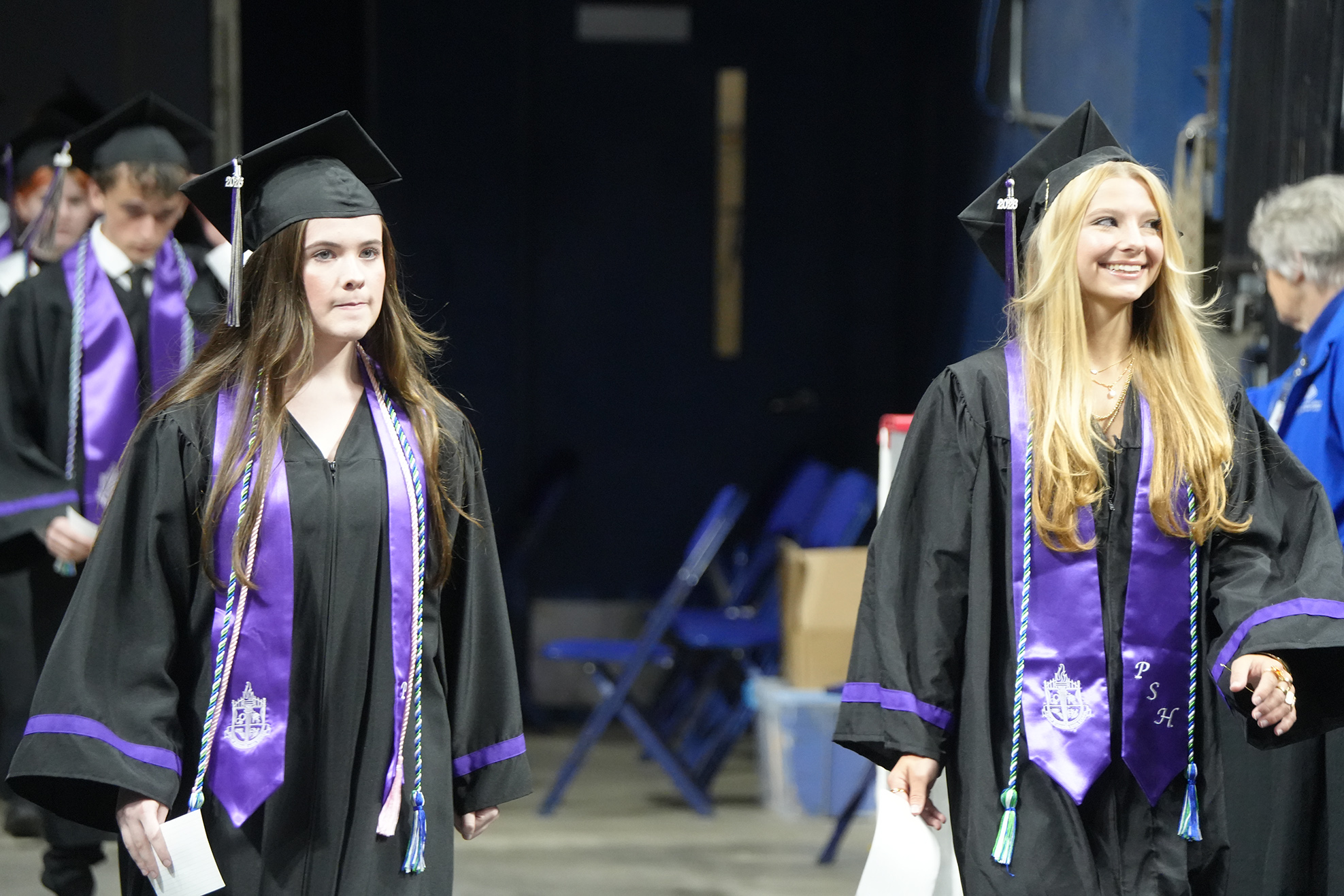 Two women in graduation gowns, one with blonde hair and the other with dark hair, walk forward.