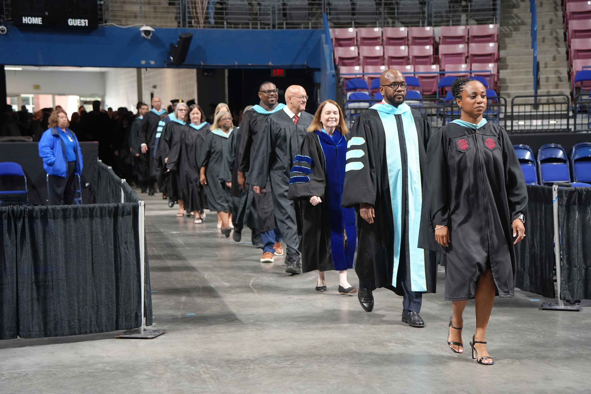 People in academic attire walk down an aisle in a large, empty auditorium with rows of blue chairs.