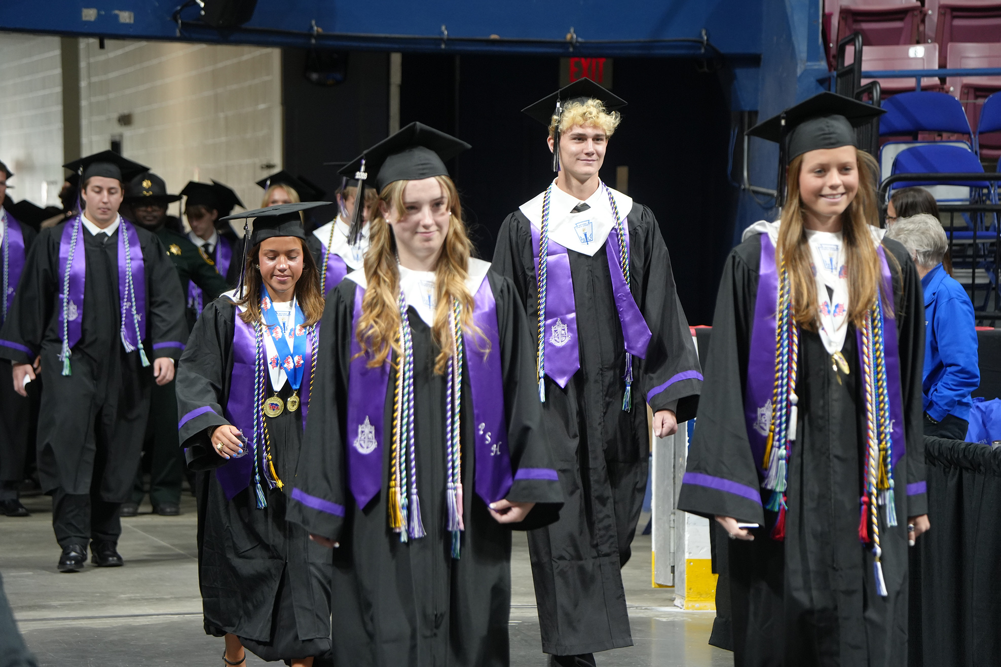 Graduates in black gowns and caps with purple stoles walk down a hall with empty seats.
