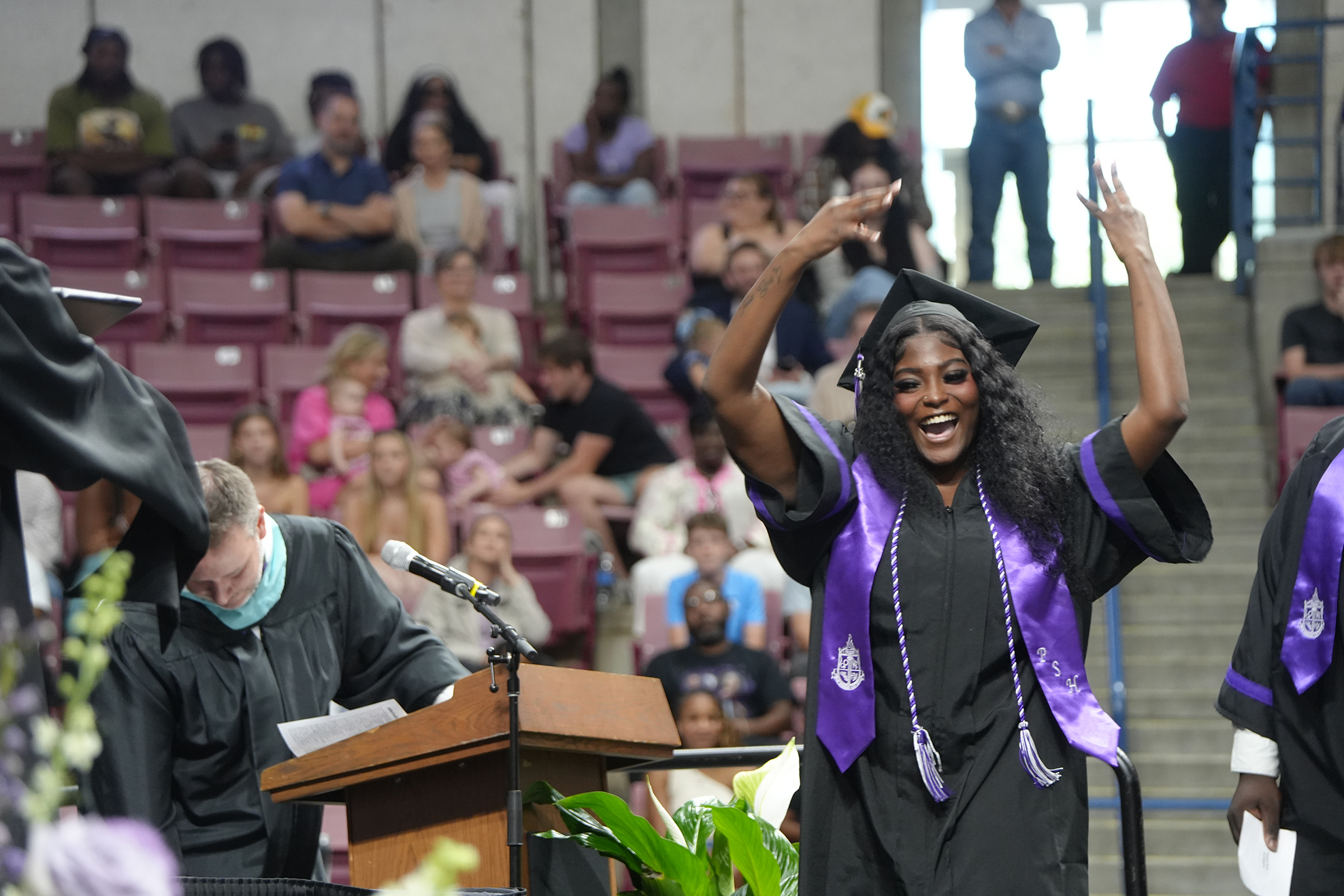 A woman in a graduation gown stands on stage with both arms raised, smiling. Behind her, a person speaks into a microphone.