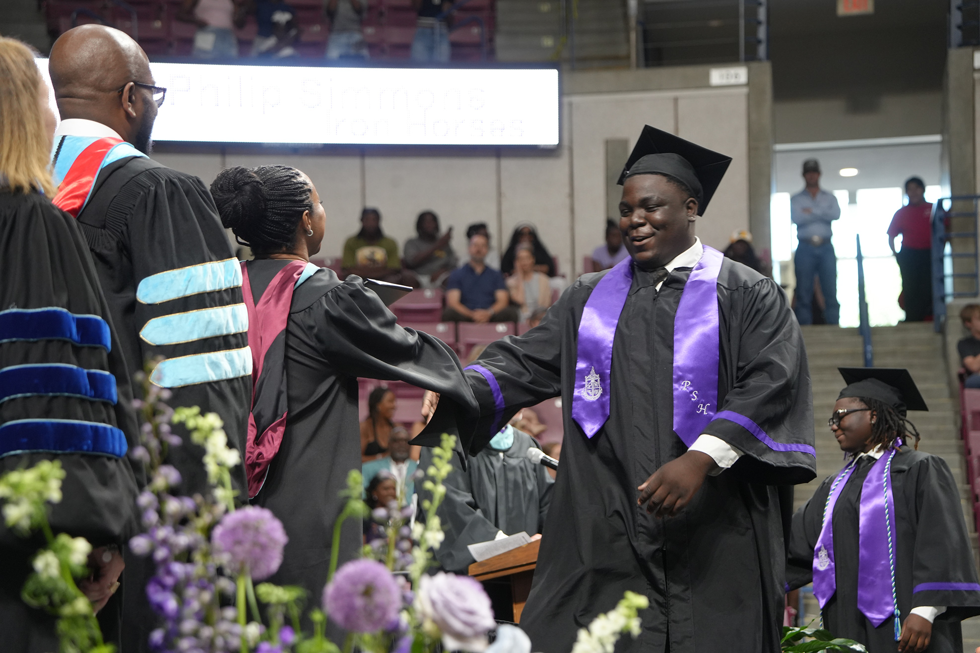A man in a graduation gown stands next to a woman in the foreground. Behind them, a large crowd of people sits on bleachers.